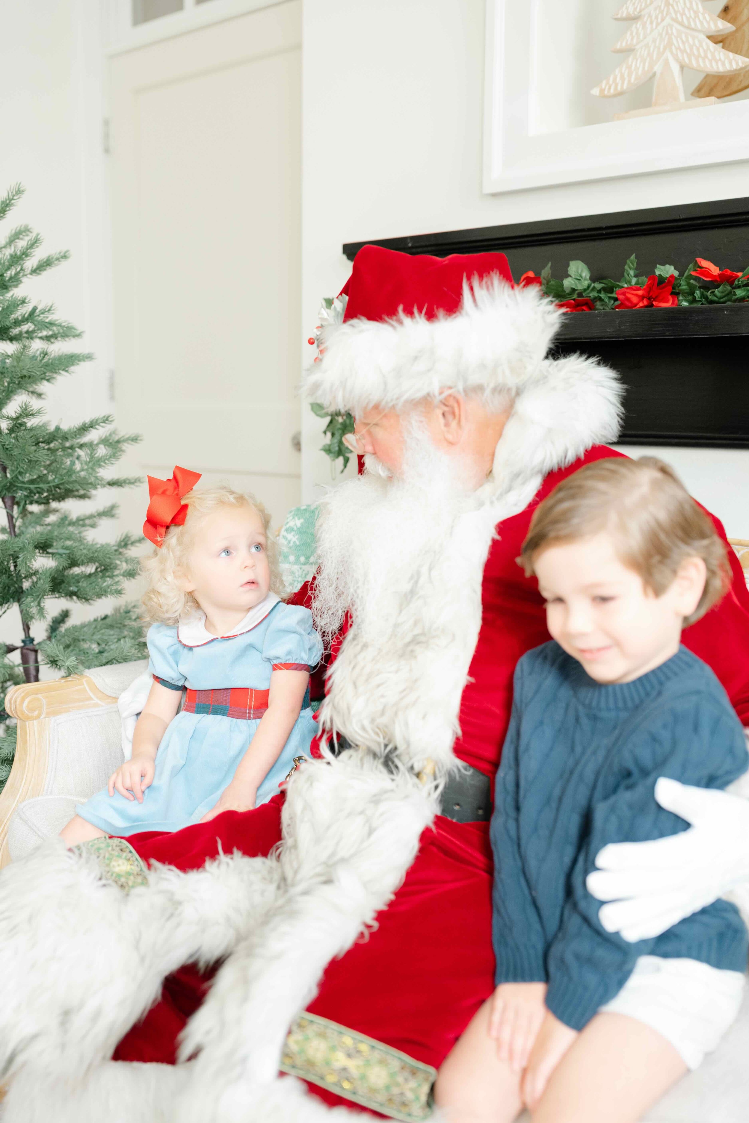 Little boy and girl sitting next to Santa and girl is looking up at Santa's eyes wondering when she will stop believing in Santa by Kelly Bullock Photography in Raleigh, NC.