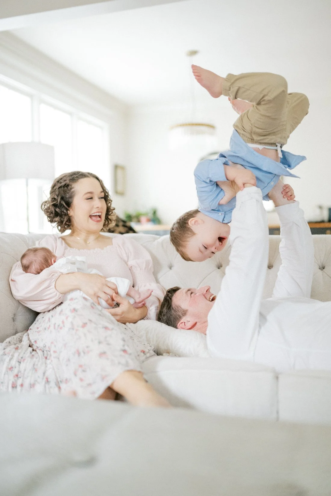 Natural family photos of a dad lifting his son upside down while mom cuddles their newborn on the couch during an in-home lifestyle session.