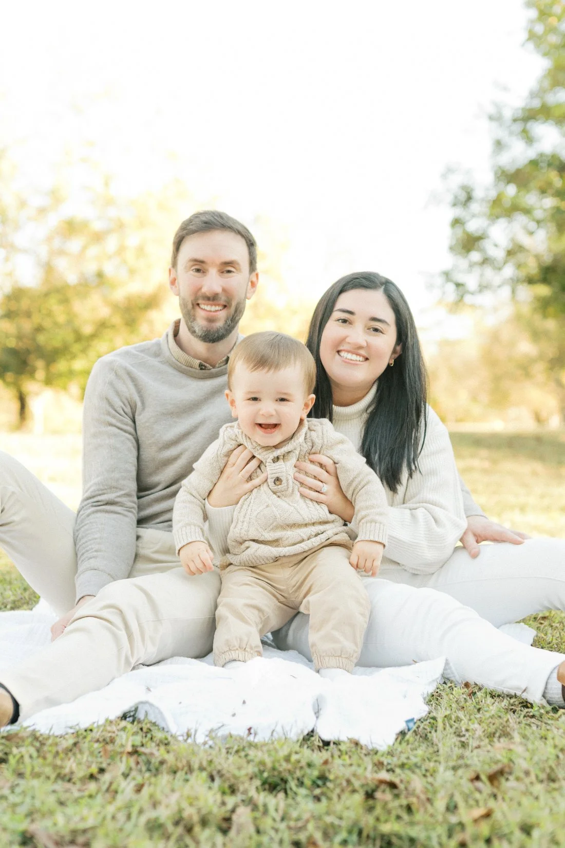 Smiling parents sitting on a blanket with their baby during soft, neutral toned spring family photos in Raleigh NC.