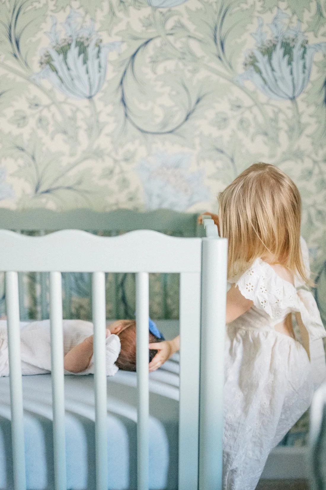 Young big sister gently touching her newborn sibling while the baby rests in a crib during an at-home session.