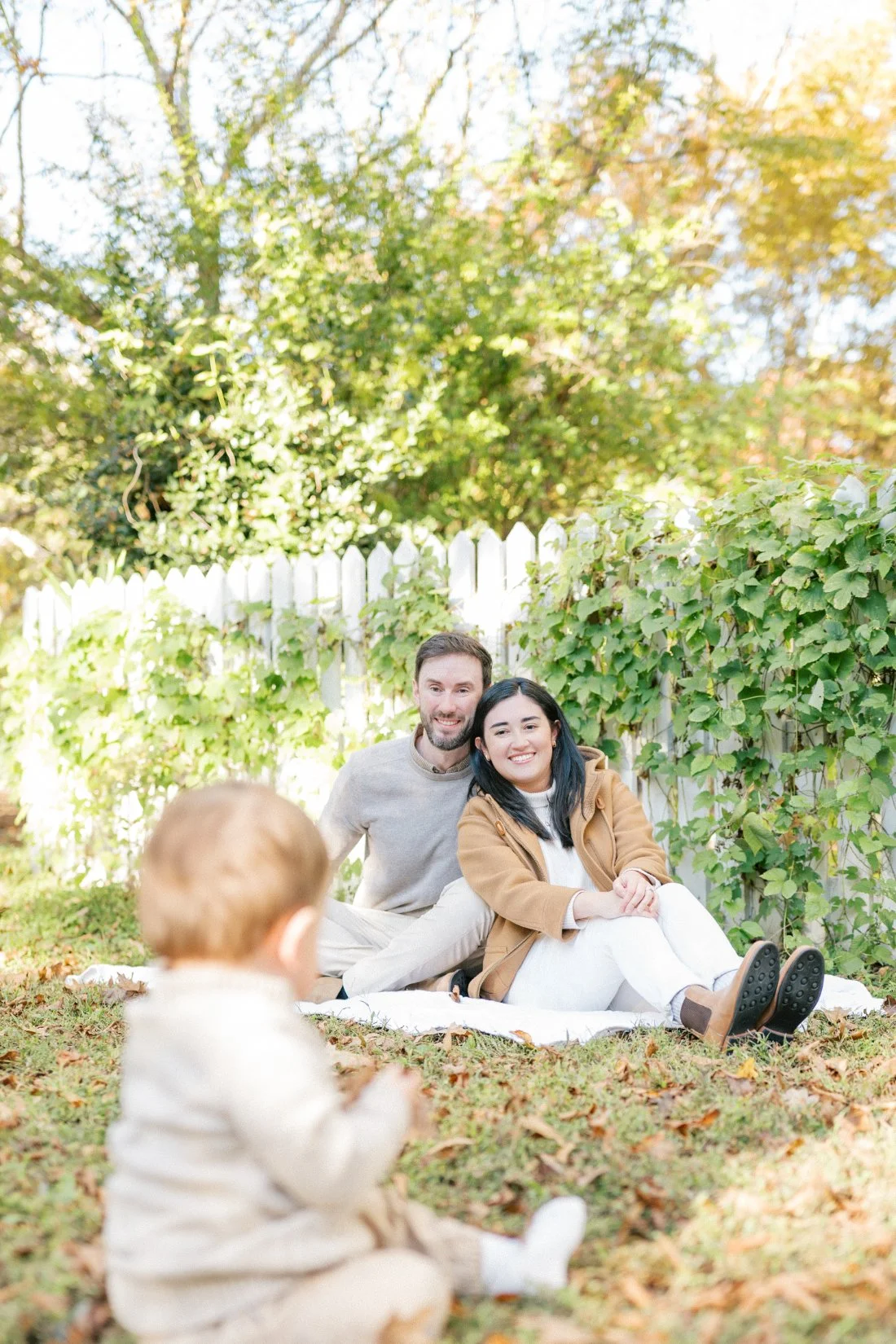 Fall family photos in Raleigh featuring parents smiling on a backyard blanket while their baby plays in the foreground.
