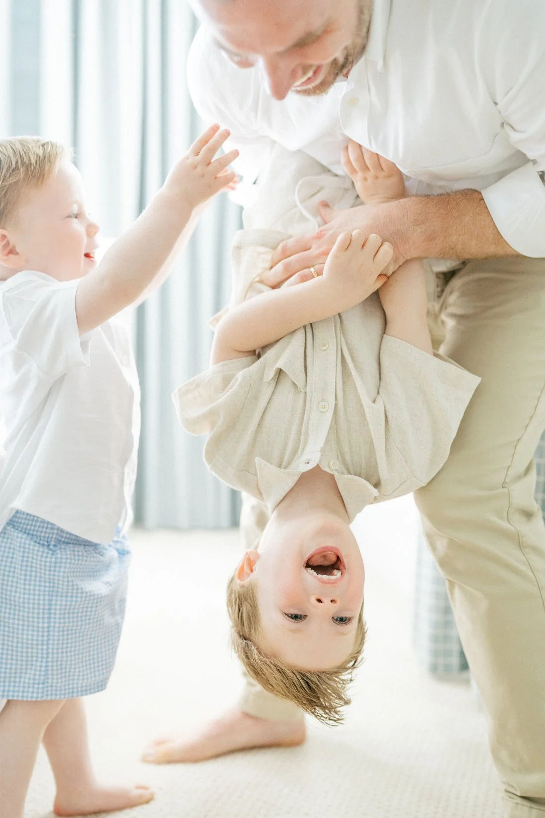 Natural family photos of a dad holding his laughing son upside down while his younger brother reaches to play during an in-home family session.