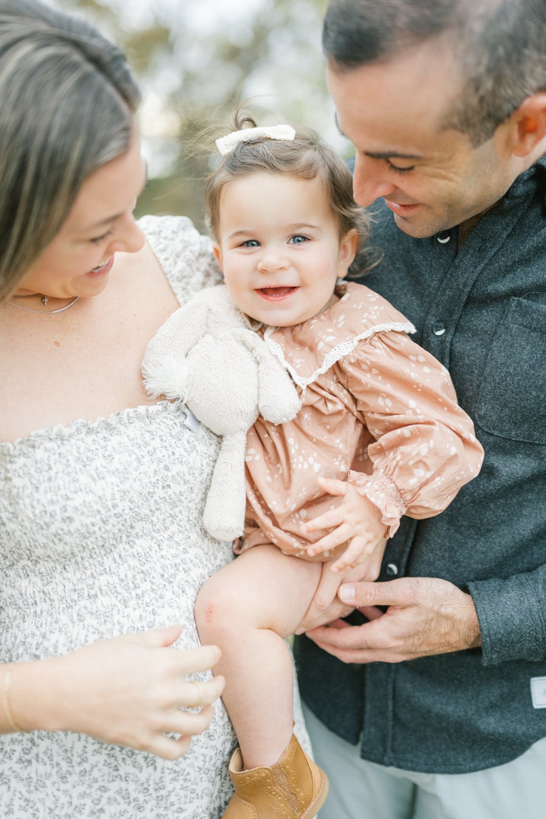 Mom and dad smiling at their baby girl as she holds her favorite stuffed bunny during a relaxed outdoor family session.