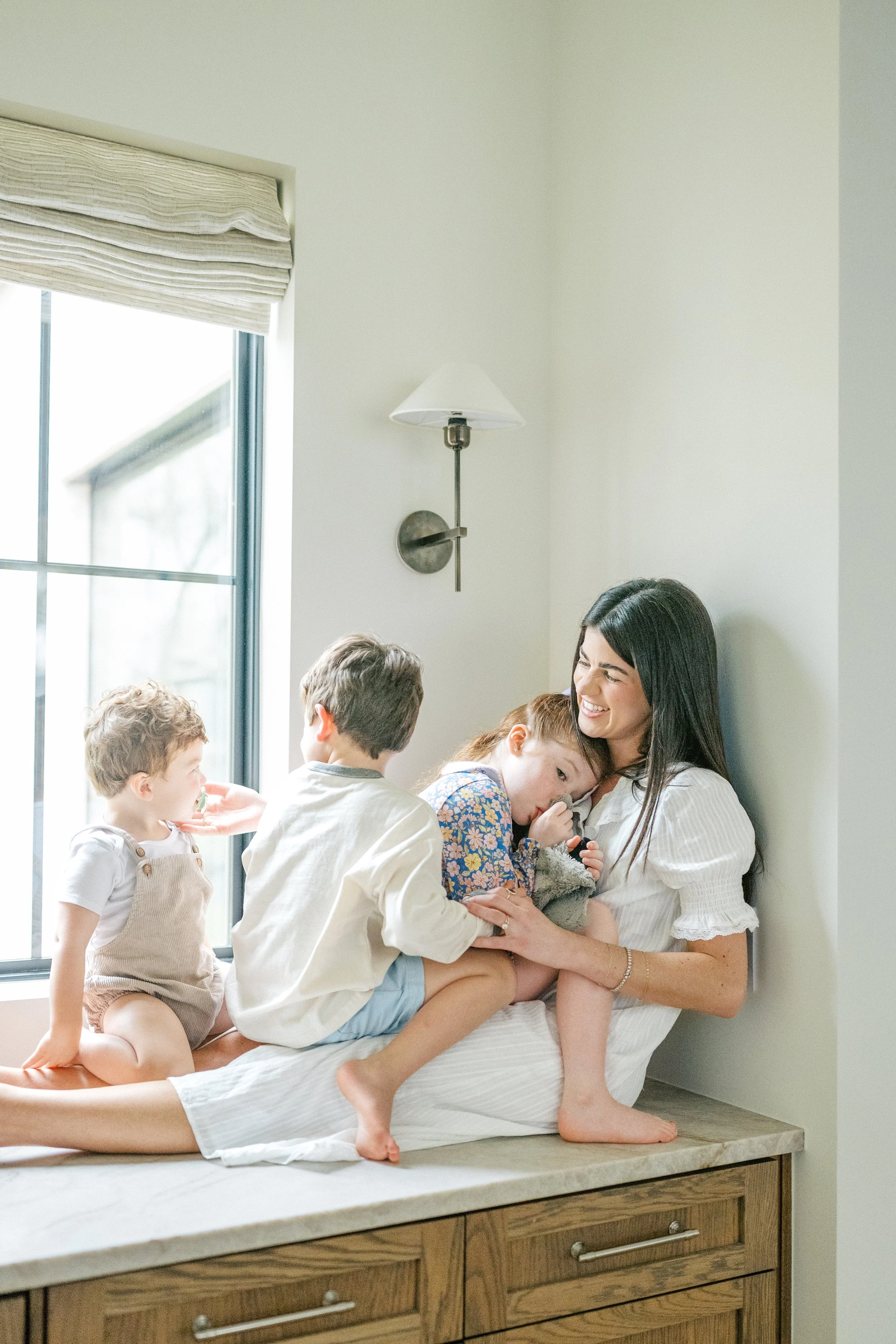 Mom sitting by a window with her three kids on her lap during an in-home family session in Austin, Texas.