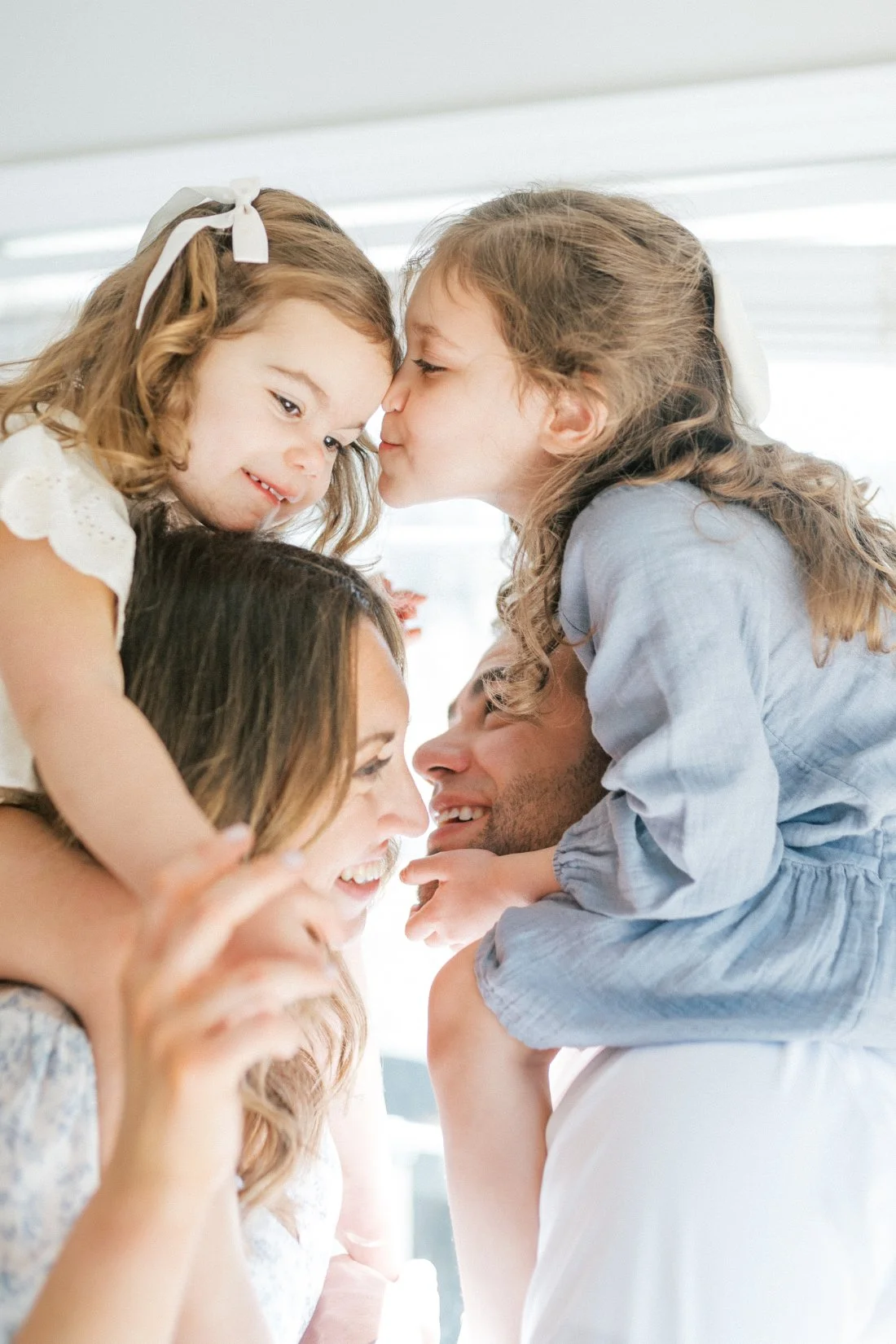 A joyful close-up of two young sisters sharing a kiss while sitting on their parents’ shoulders, all smiling and snuggled together — capturing real love and laughter through candid family photos.