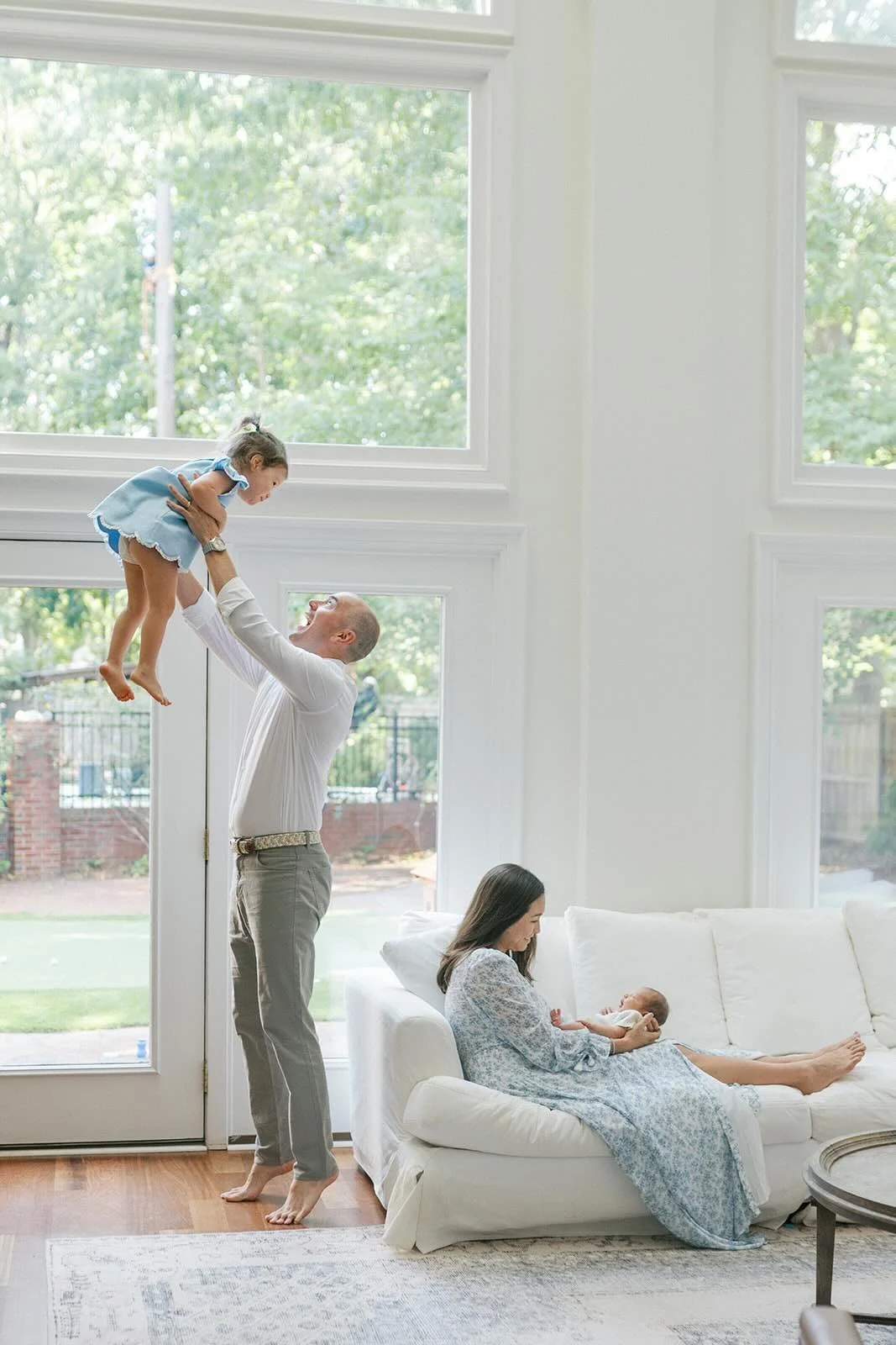 Father playing with big sister while mother holds baby boy in the natural light of the large windows - Raleigh Newborn Photographer