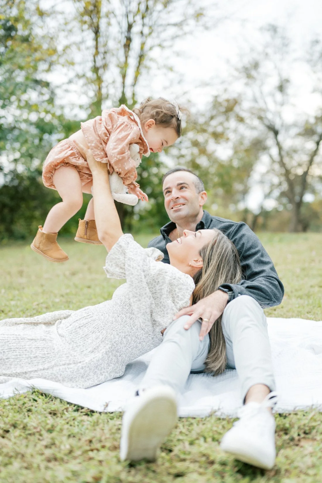 Toddler laughing while being lifted into the air by her mother as dad watches during an outdoor family photography session