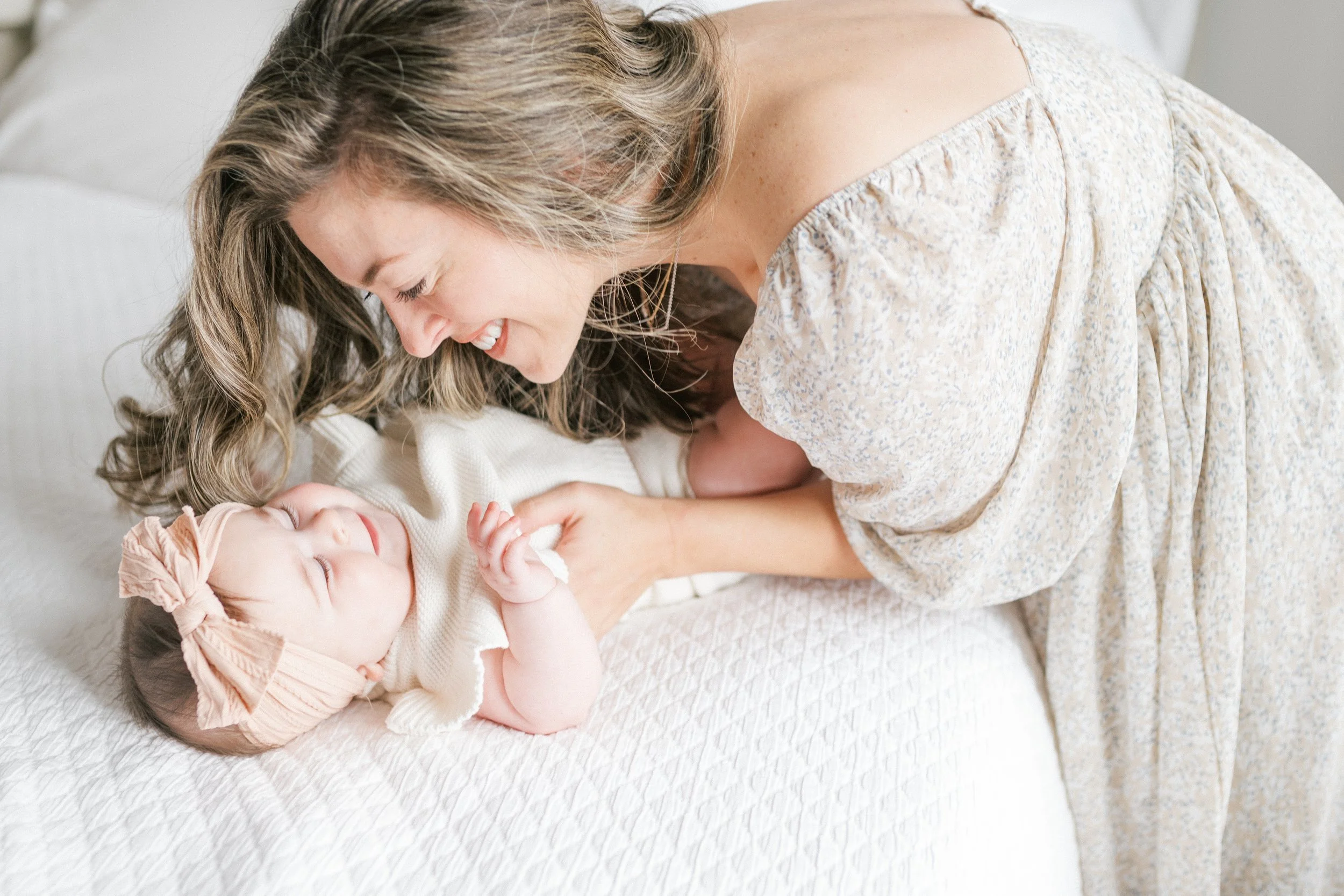 Tender mom smiling nose-to-nose with her baby on a white bed in soft neutral tones—how to prepare for an in-home photography session.