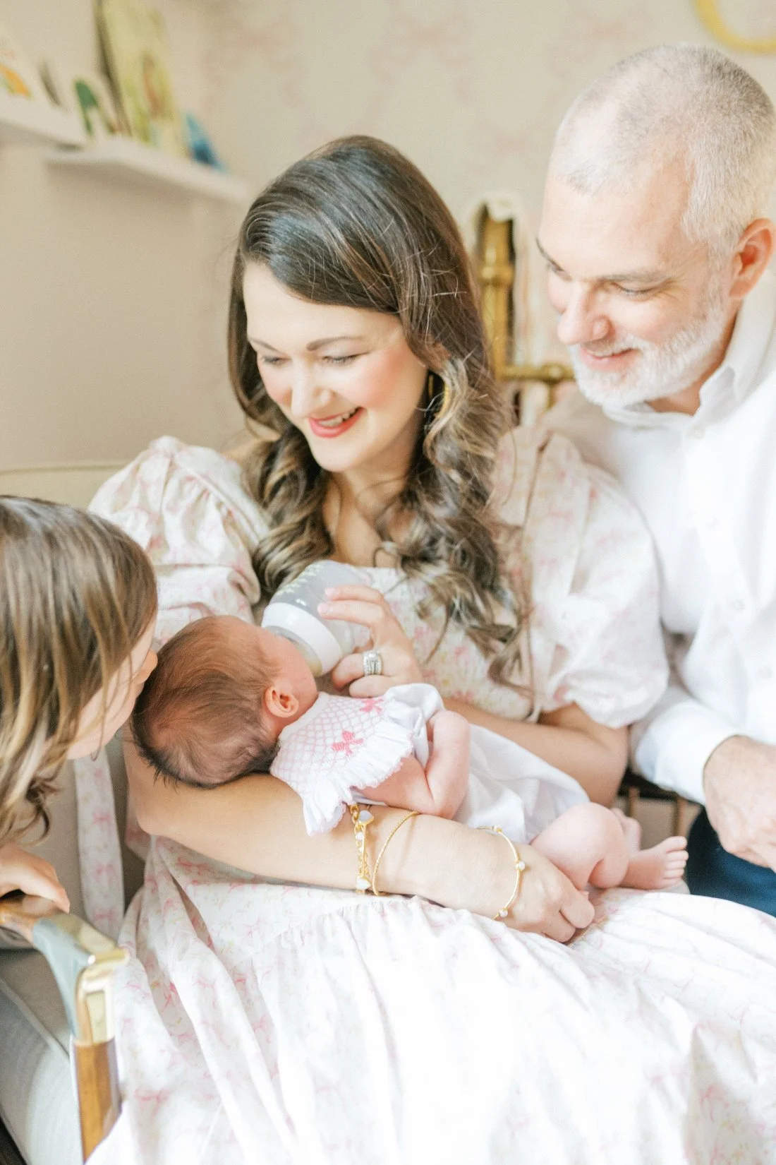 A sweet newborn feeding moment as a mother bottle-feeds her baby while smiling, surrounded by dad and big sister — an intimate scene that highlights the beauty of candid family photos at home.