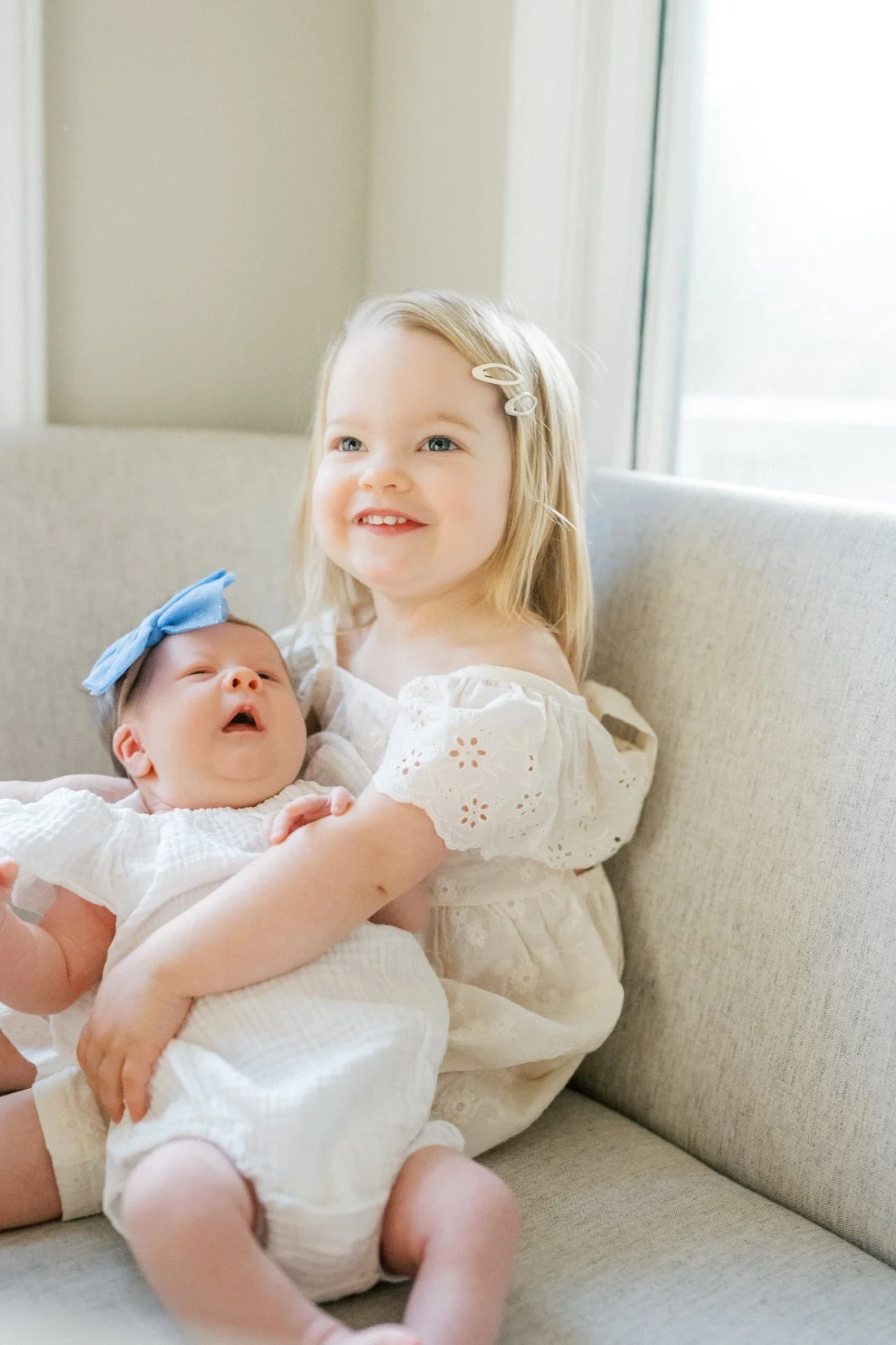 Toddler holding newborn sibling on couch during in-home milestone checklist session with soft natural light
