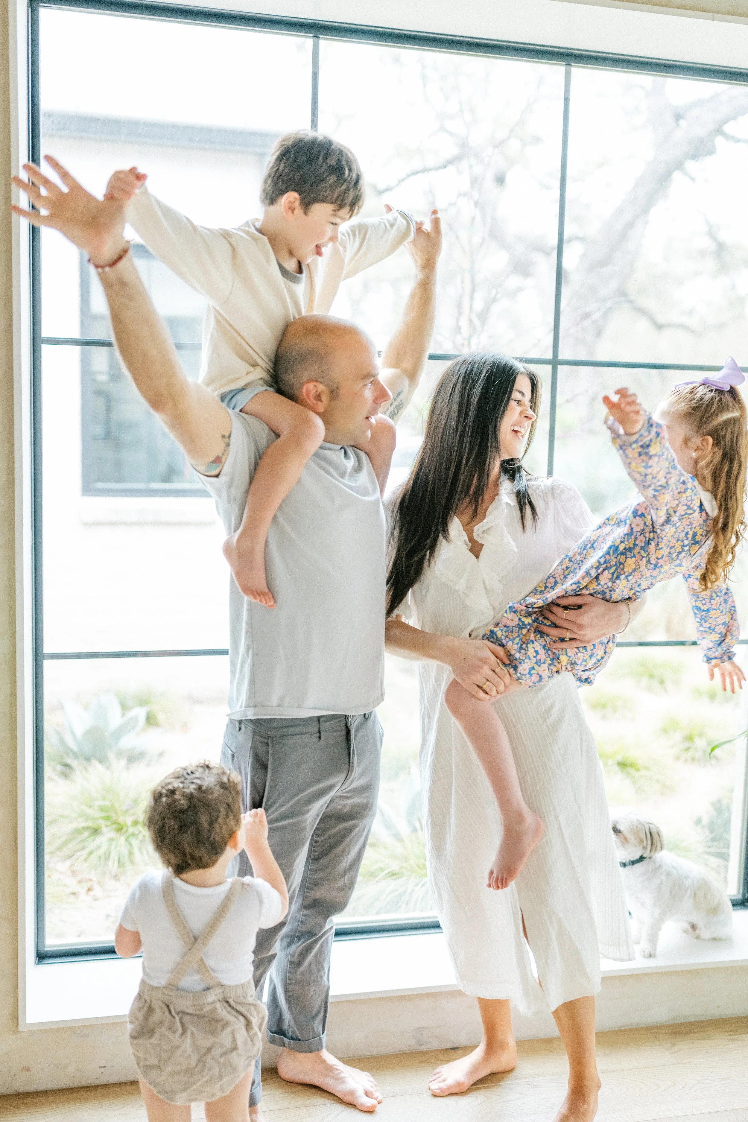Dad holding boy on his shoulders and mom holding little girl on her arms, standing by a window during an family session in Austin, Texas.