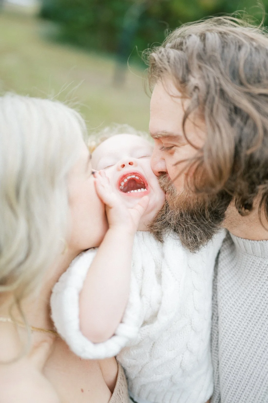 Parents kissing their giggling toddler on the cheeks during a joyful outdoor family photography session.