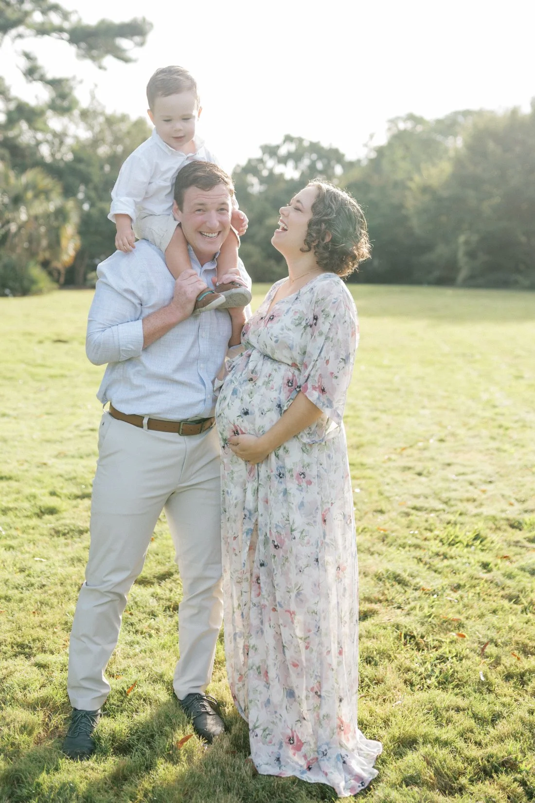 Smiling family outdoors with toddler on father’s shoulders and expecting mother in flowy floral dress