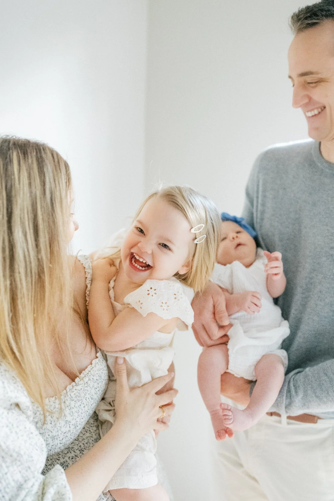 Newborn sibling photos of a smiling toddler with her mom while dad holds her new baby sister.