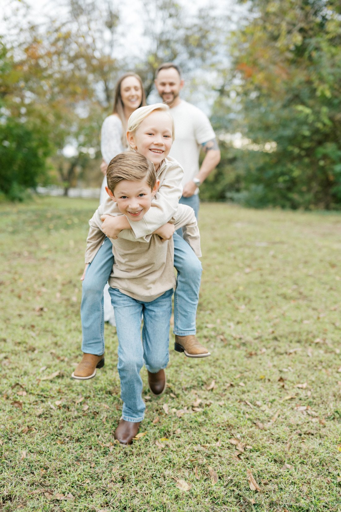 Two brothers laughing during fall family photos in Raleigh as the younger brother rides piggyback with their parents smiling behind them.