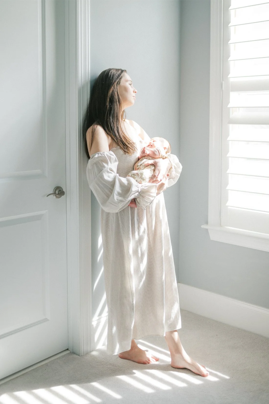 Mother holding her newborn beside a sunlit window during a quiet in-home newborn session.