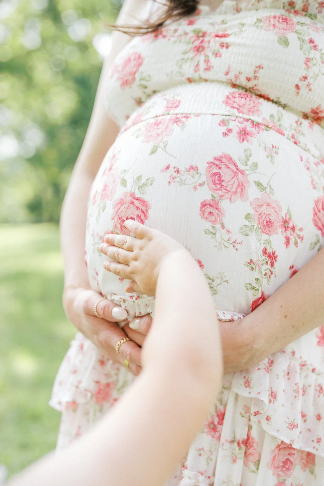 Close up of pregnant belly in floral dress with child’s hand resting on bump during spring maternity photos