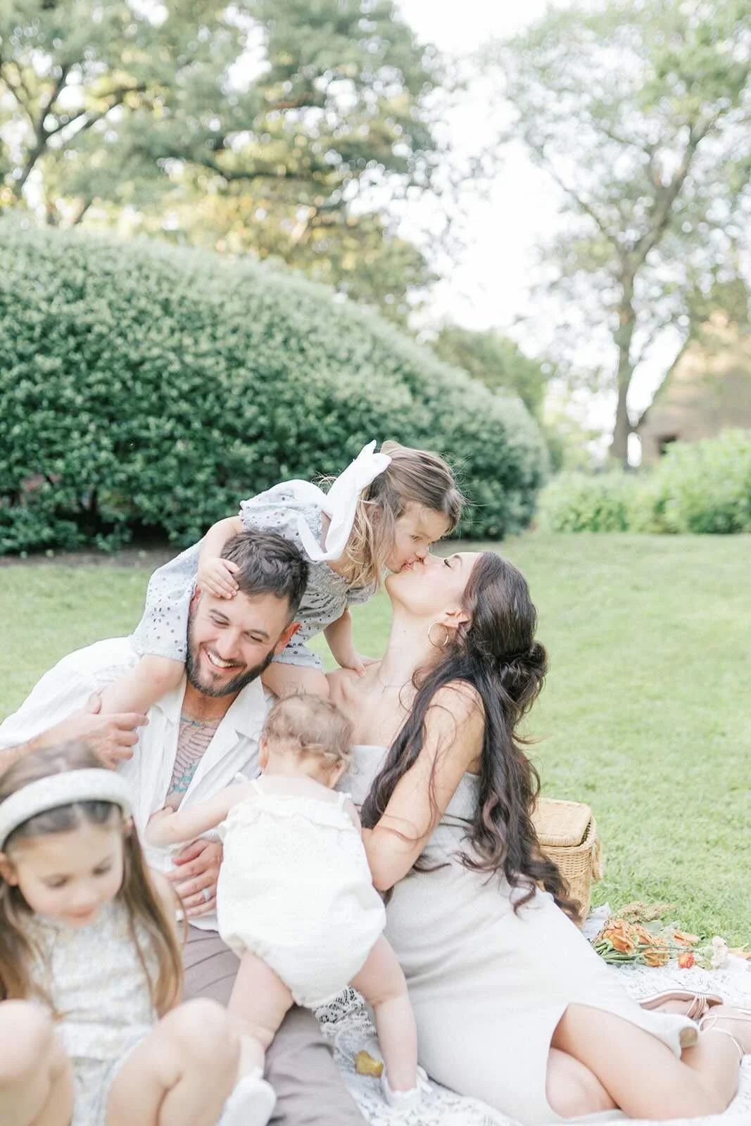 Family sitting on a blanket outside while Mom gives daughter a kiss while on Dads shoulders - Raleigh Newborn Photographer