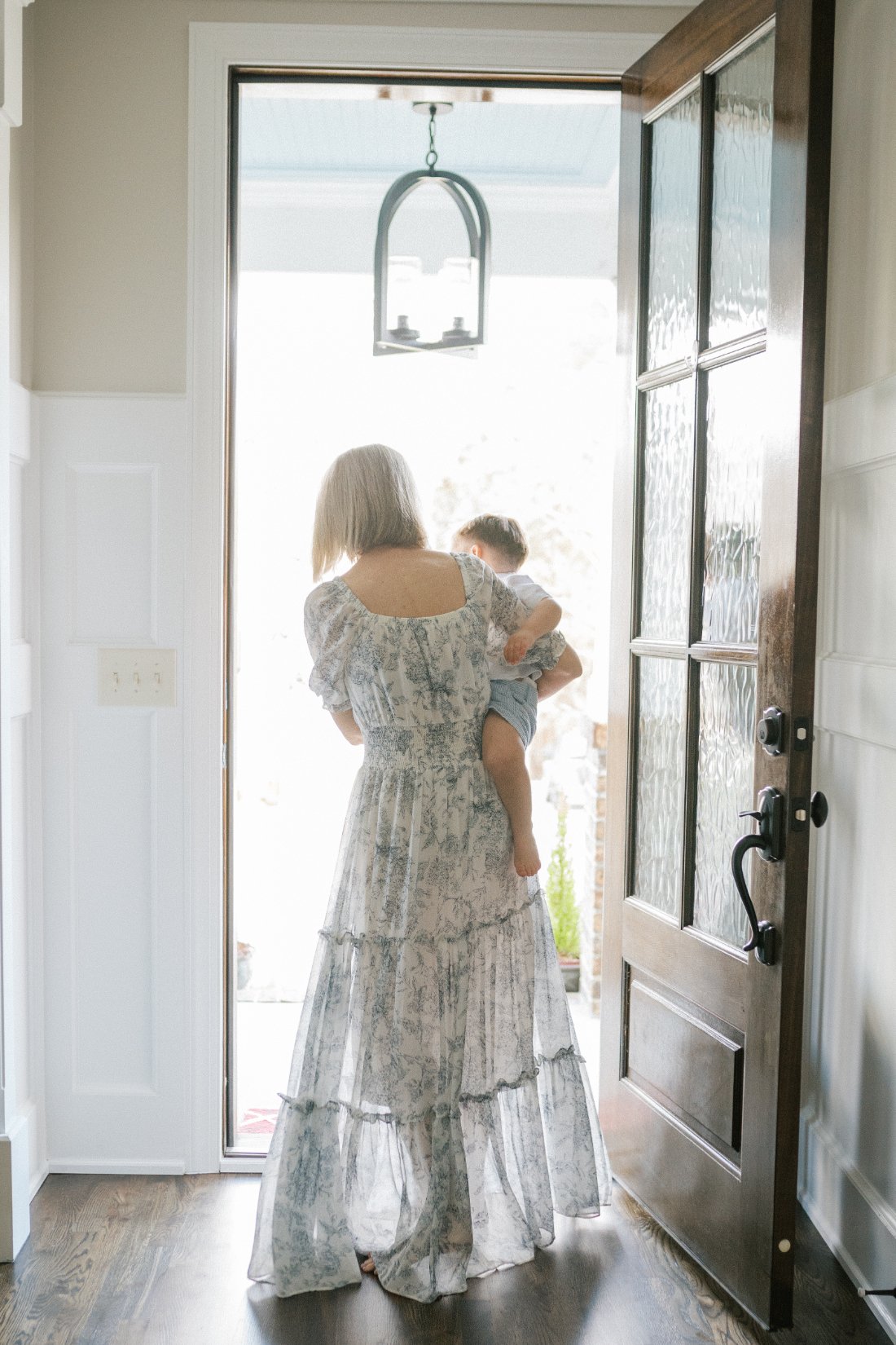 Grandparent photoshoot scene of a grandmother carrying her young grandchild while walking through a bright doorway, capturing a quiet and tender in home moment.