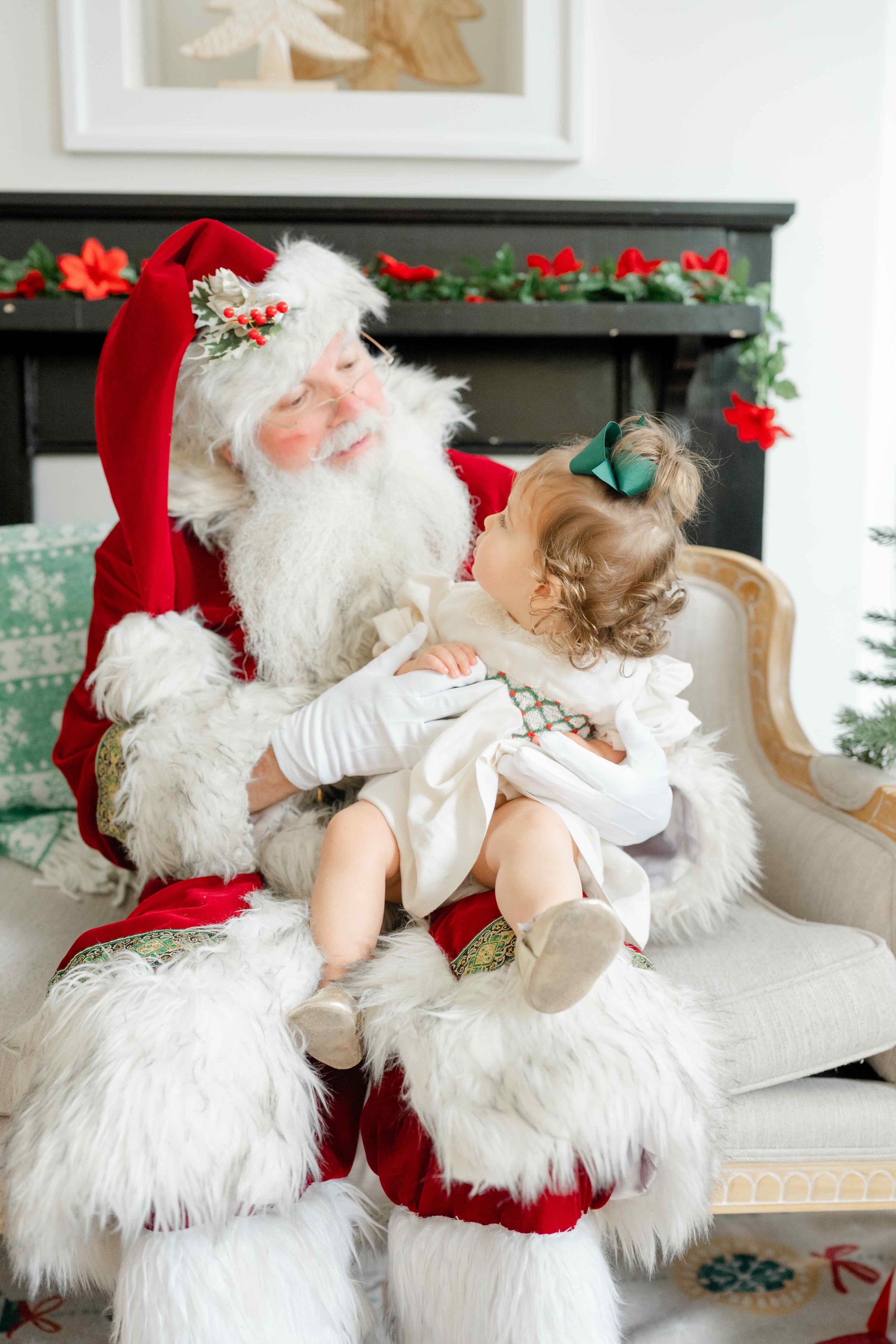 Little girl sitting in Santa's lap and they're looking at eachother wondering when she will stop believing in Santa by Kelly Bullock Photography in Raleigh, NC.