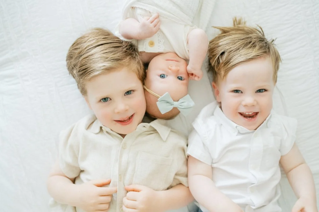 Three siblings lying together on a bed, including a newborn baby between two smiling brothers during a Raleigh photographer family session