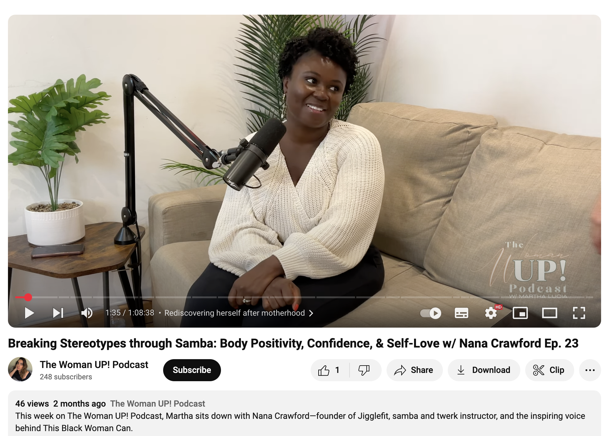 Nana Crawford, a Black woman with short curly hair, in a cream sweater, sitting on a beige couch, speaking into a black microphone during a podcast interview, with green plants and a small wooden side table in the background.