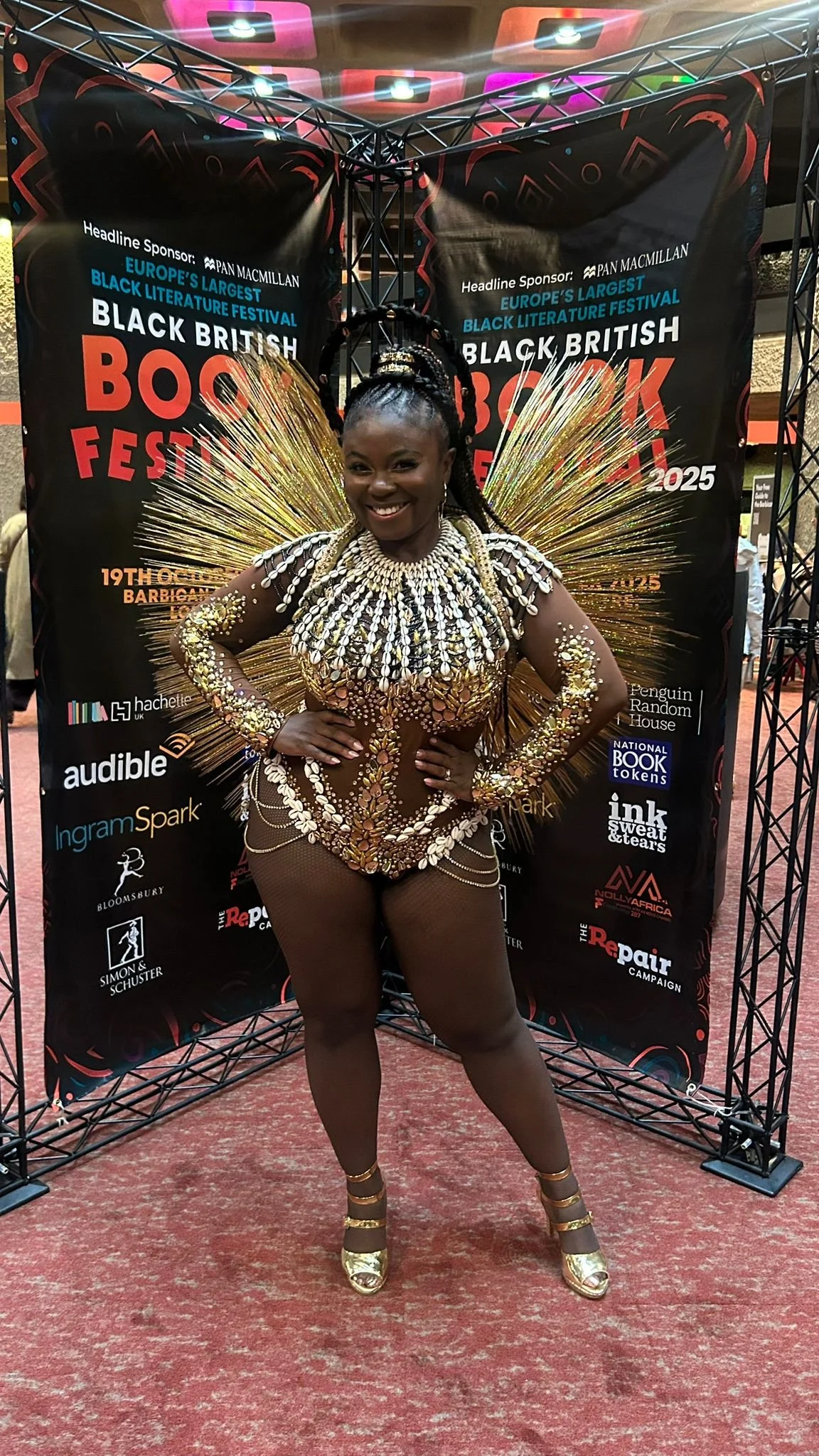 A woman dressed in an elaborate, gold and white costume with fairy wings, posing in front of a backdrop at the Black British Book Festival.