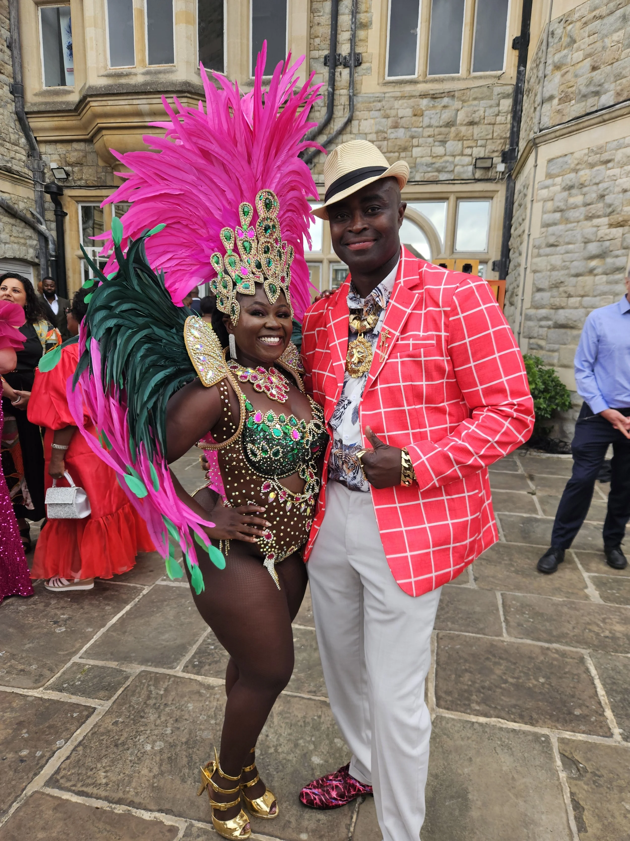 A woman in a colorful carnival costume with feathers and jewelry and a man in a red checked blazer and beige hat posing together outdoors.