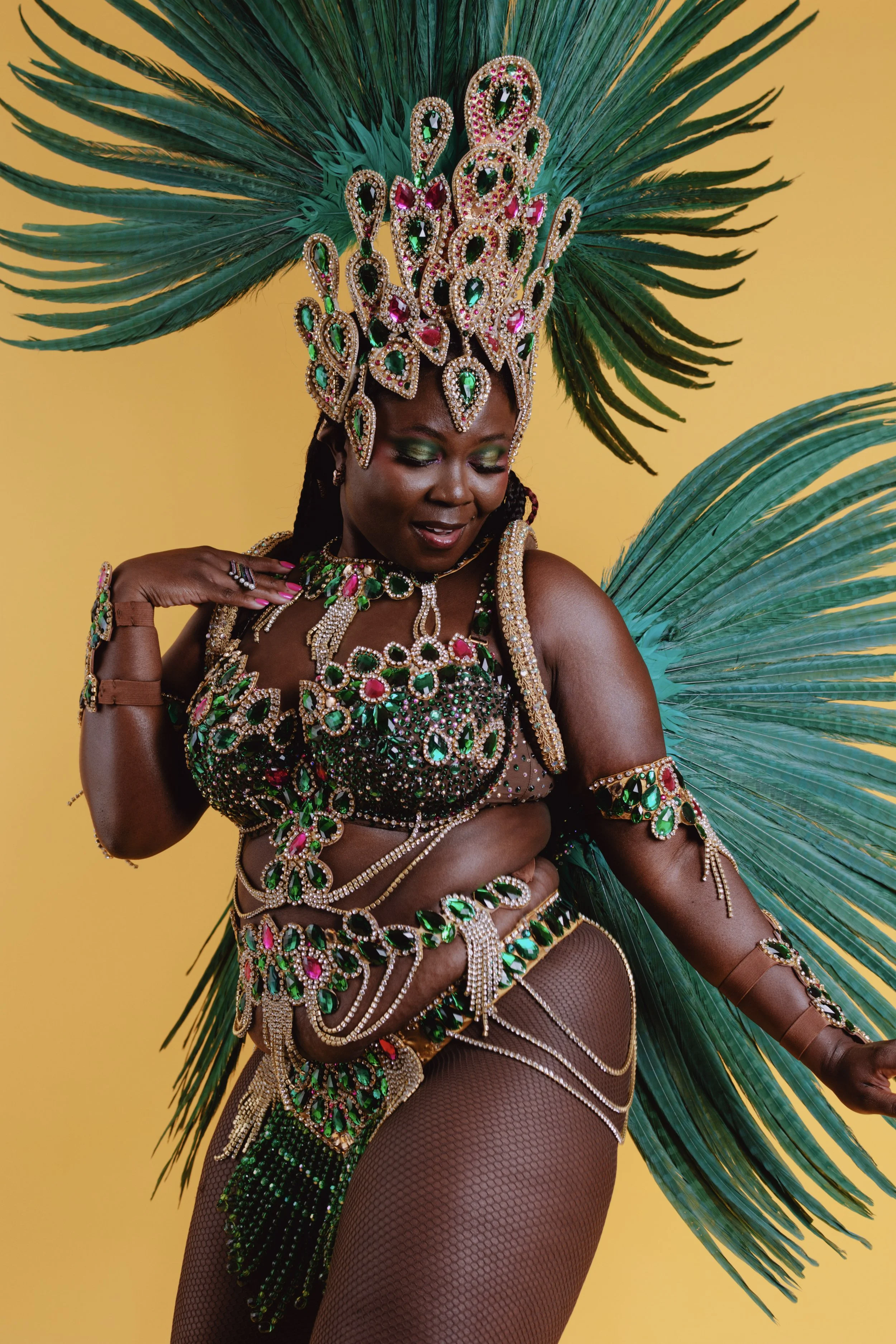 Person wearing a colorful carnival costume with green feathers and gemstone embellishments on a yellow background.