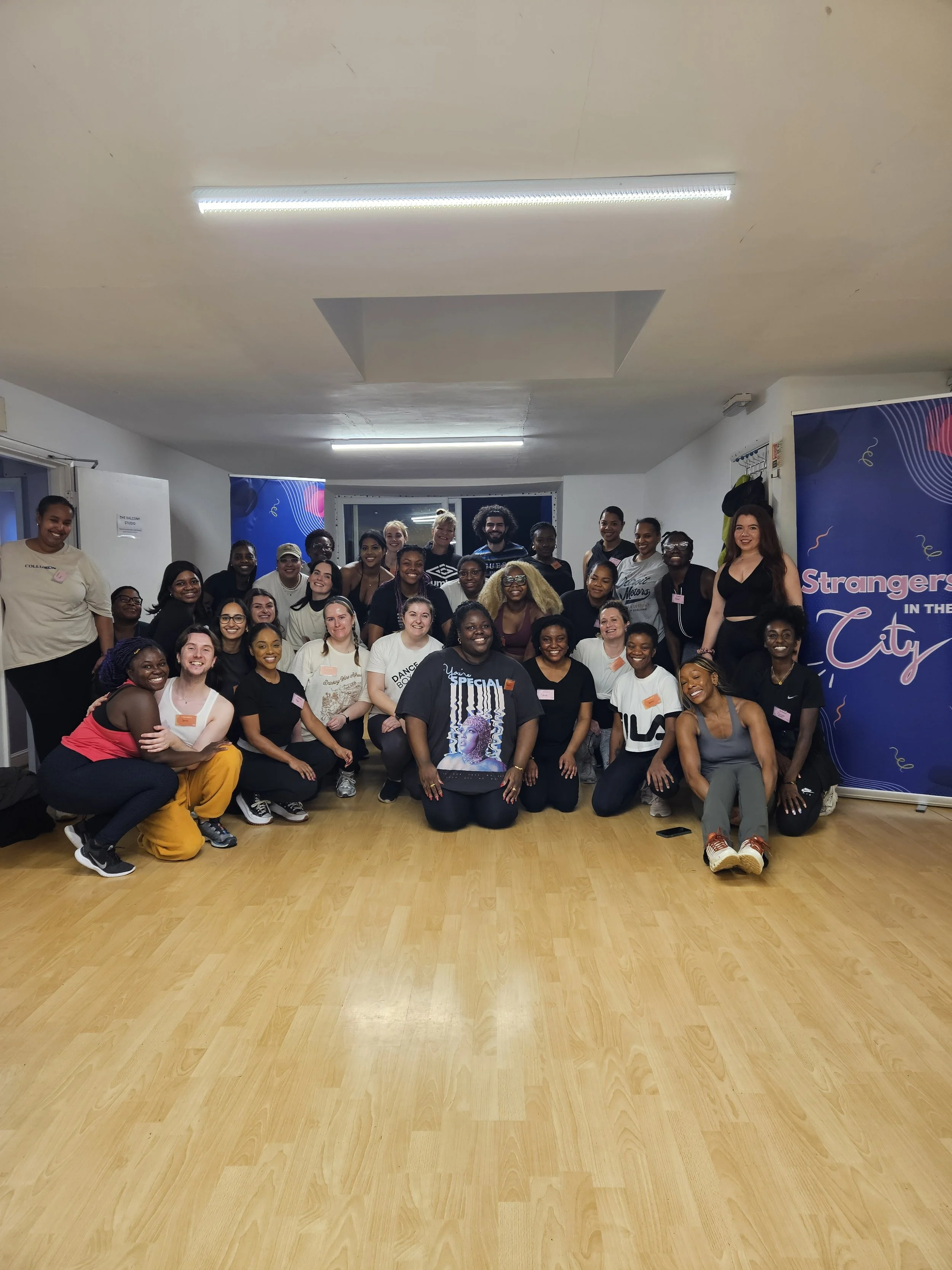 Group of diverse women posing indoors in front of a blue banner that says 'Strangers in the City' with a wooden floor and white walls.
