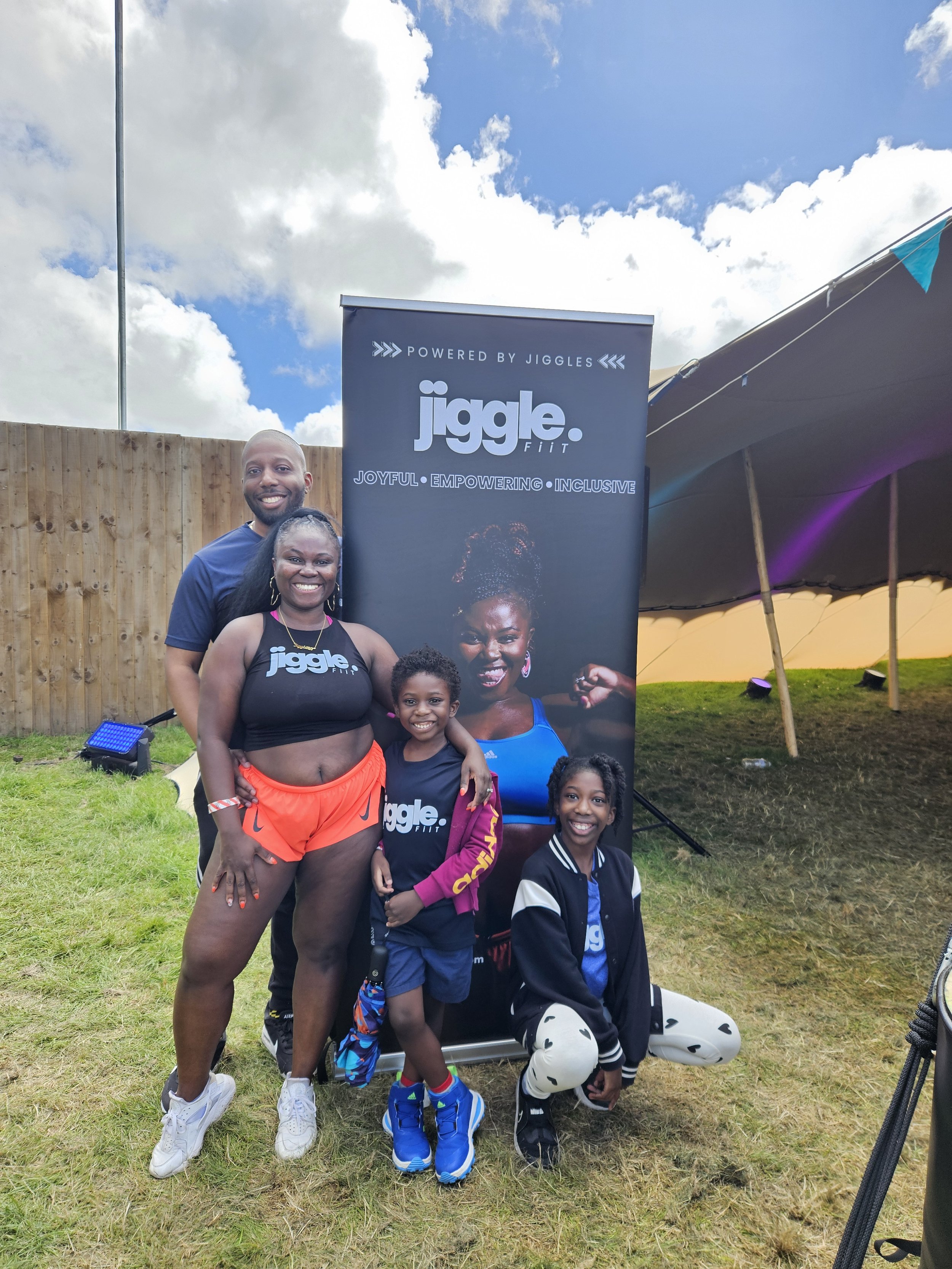Group of four children and two adults standing outdoors at a fitness event, posing in front of a Jiggle Fit banner. The group is smiling and appears happy.