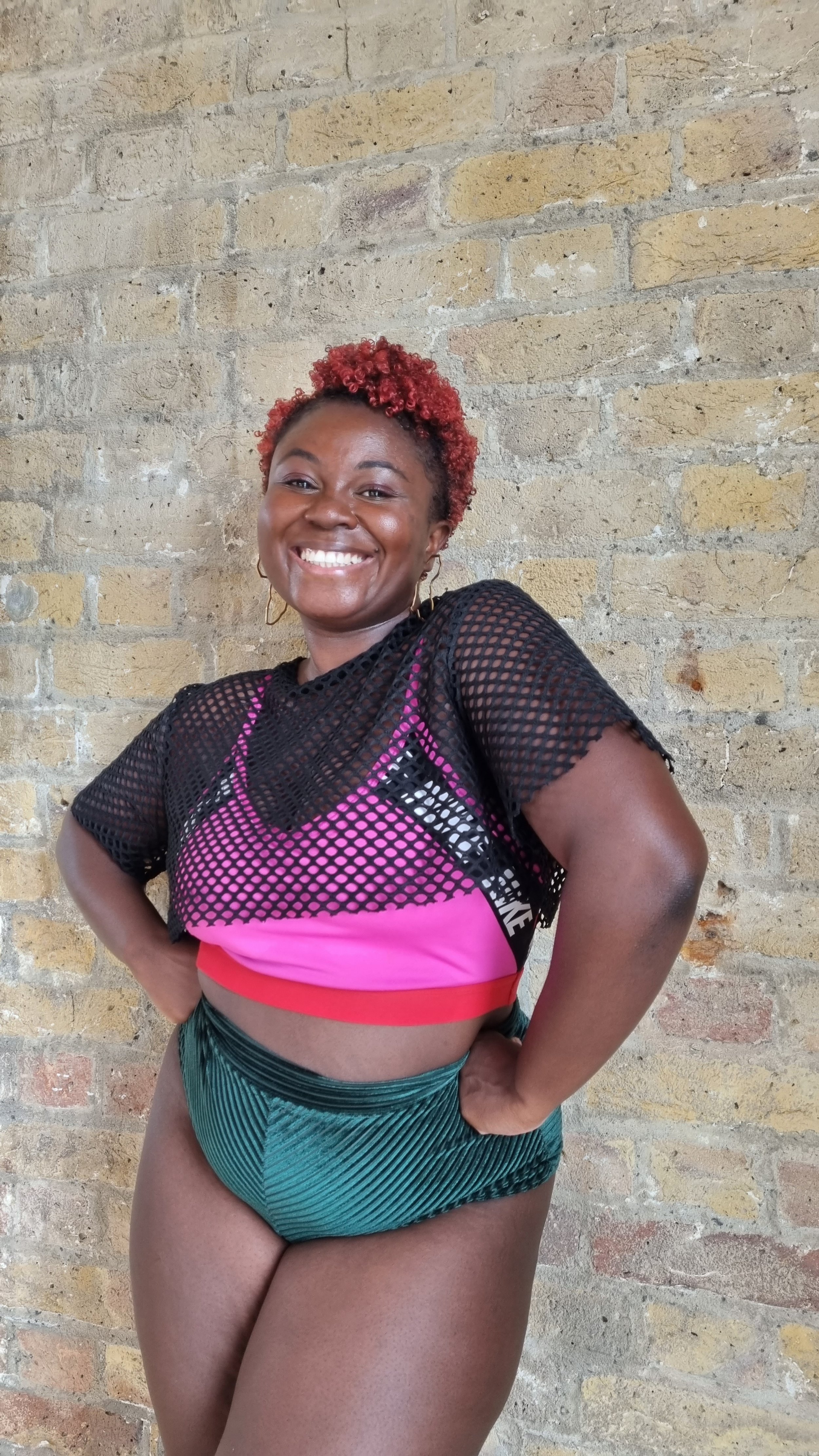 Smiling woman with short curly red hair, wearing a black mesh top over a pink sports bra and striped green shorts, standing against a brick wall.