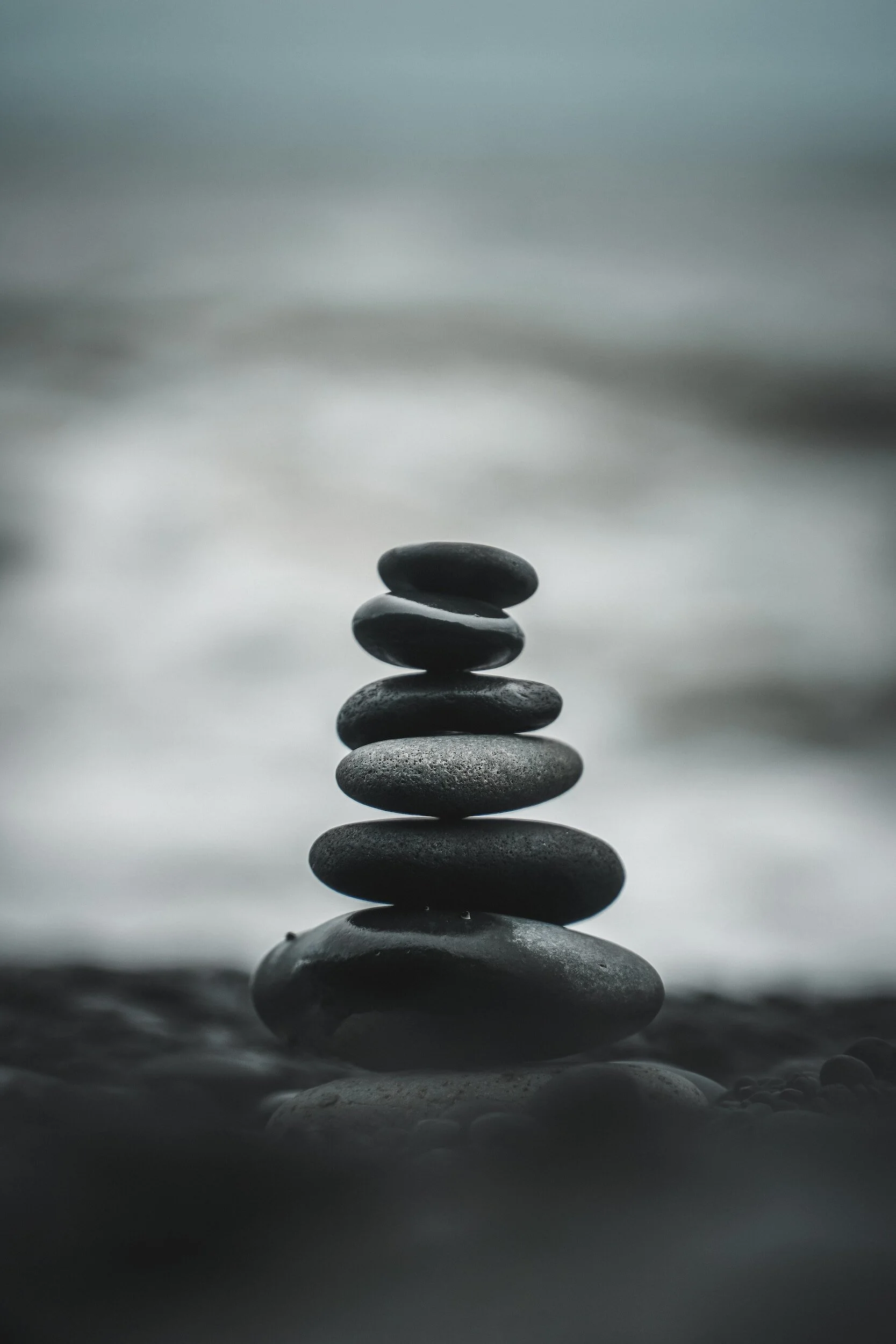 Stack of black smooth stones balanced on top of each other on a surface near water, with a blurred background