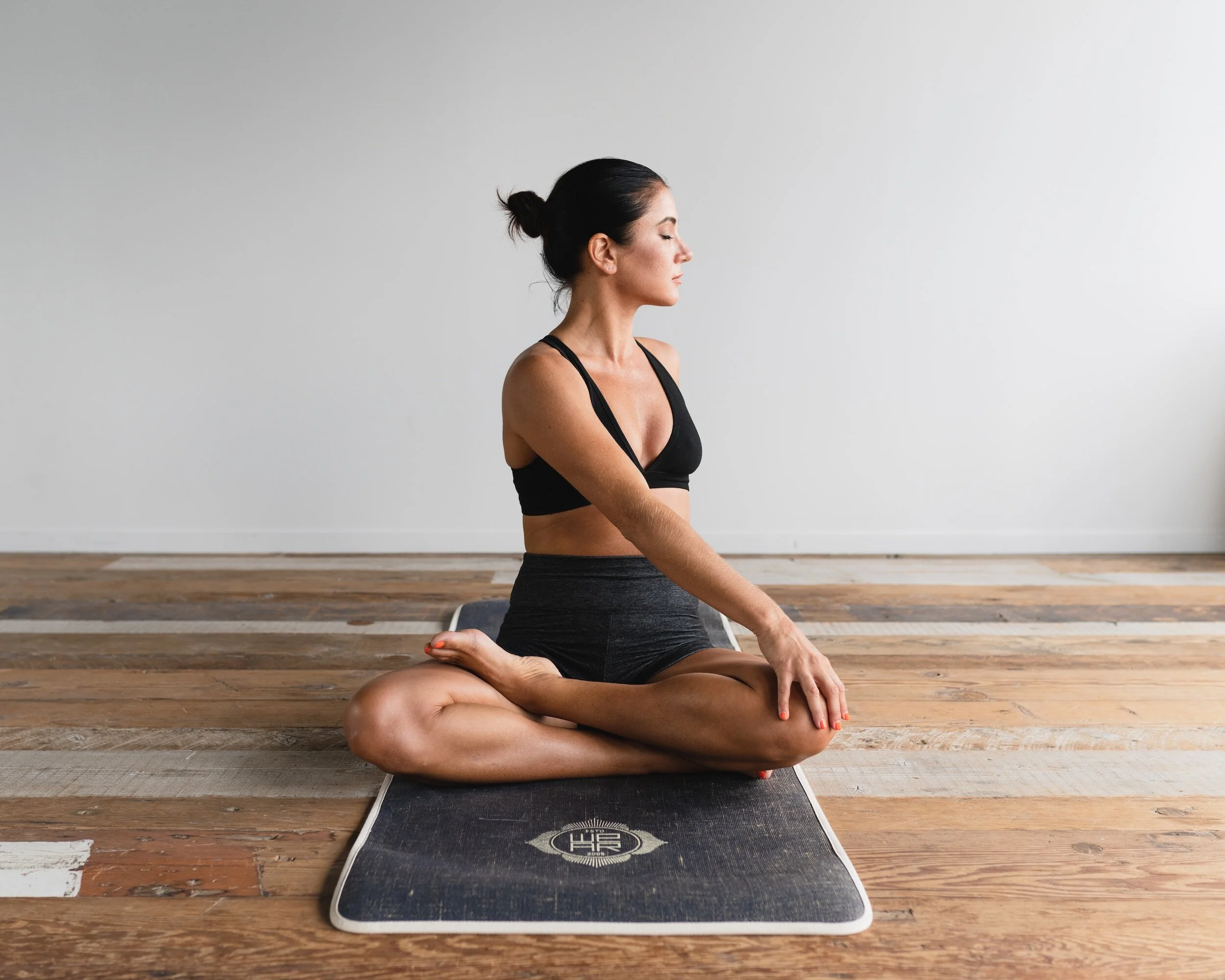 Woman practicing yoga seated in a cross-legged position on a yoga mat in a minimalist room.
