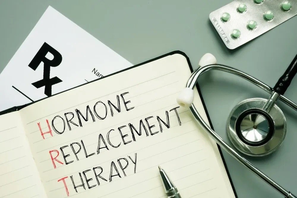 A notebook with the words "Hormone Replacement Therapy" written on it, surrounded by medical items including pills, blister packs, a stethoscope, a syringe, a small vial, and a white pill bottle on a blue background.