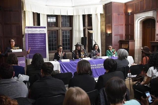 Photo of a room of people seated in chairs before a panel of four Black women, with a white woman giving an introduction at a podium to the left of the panel
