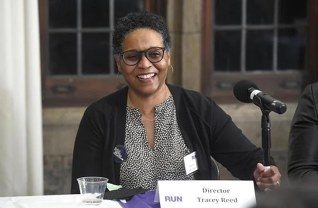 Photo of a Black woman seated at a panel table. She is wearing glasses, a black and white blouse, and a black cardigan, smiling whilst she adjusts her microphone