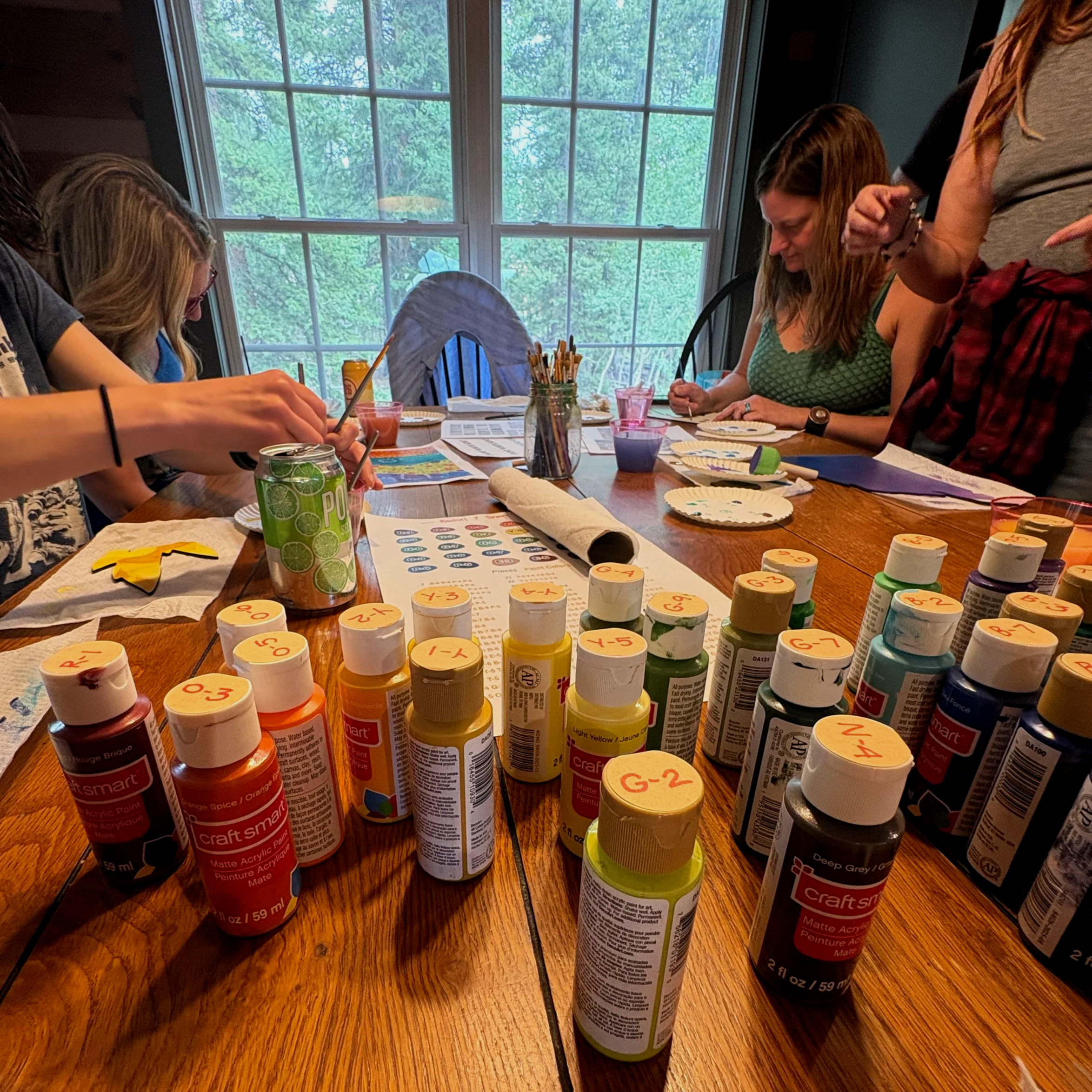A collection of brightly colored paint bottles with friends painting in the background