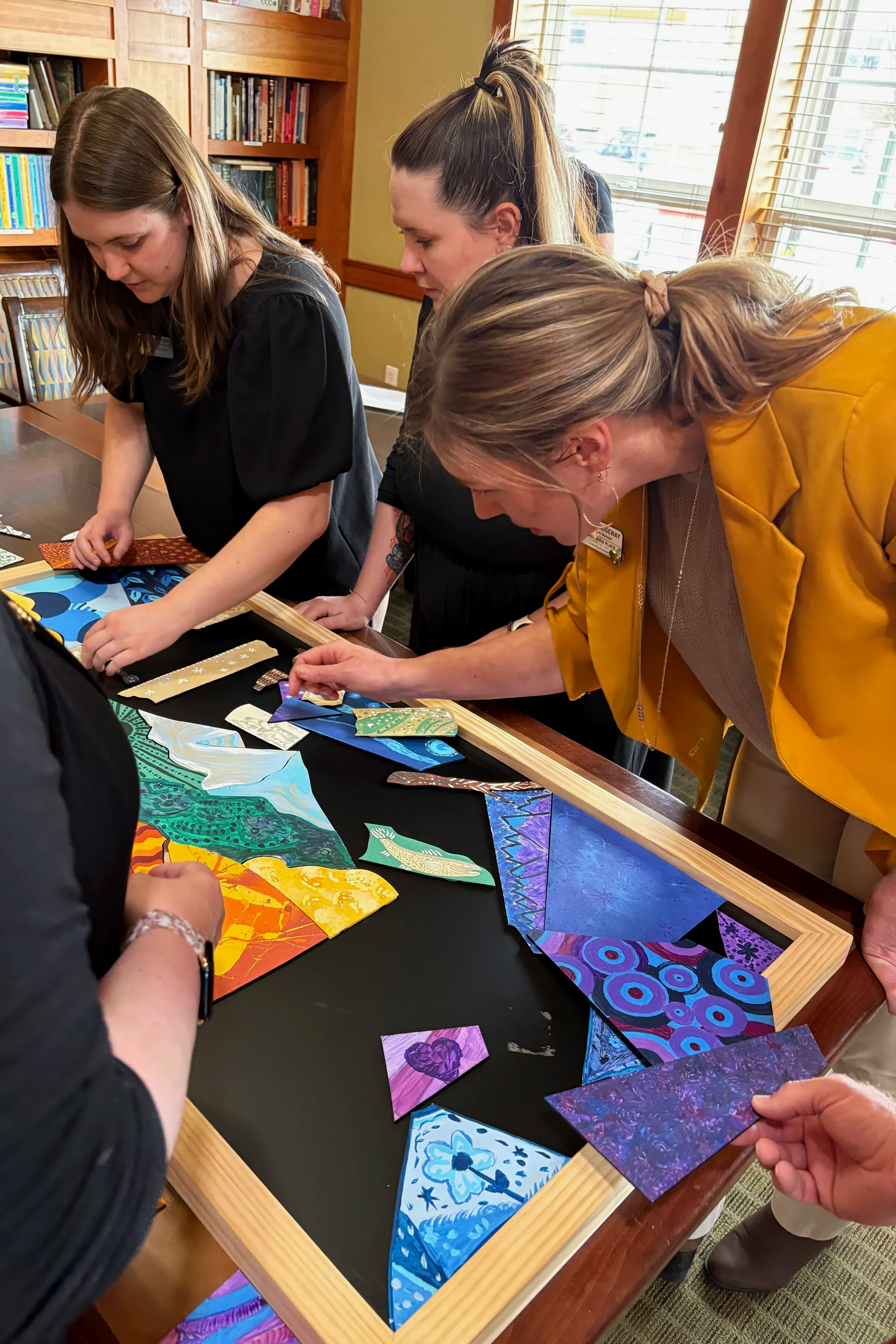 People examining colorful, patterned fabric swatches or artwork on a table inside a room with bookshelves and windows.