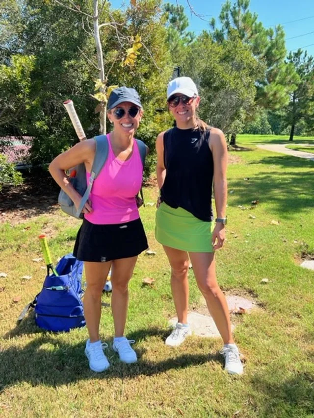 Two women in tennis outfits standing on a grassy area in a park-like setting with trees, smiling at the camera. One is holding a tennis racket over her shoulder, and a blue tennis bag is on the ground near them.