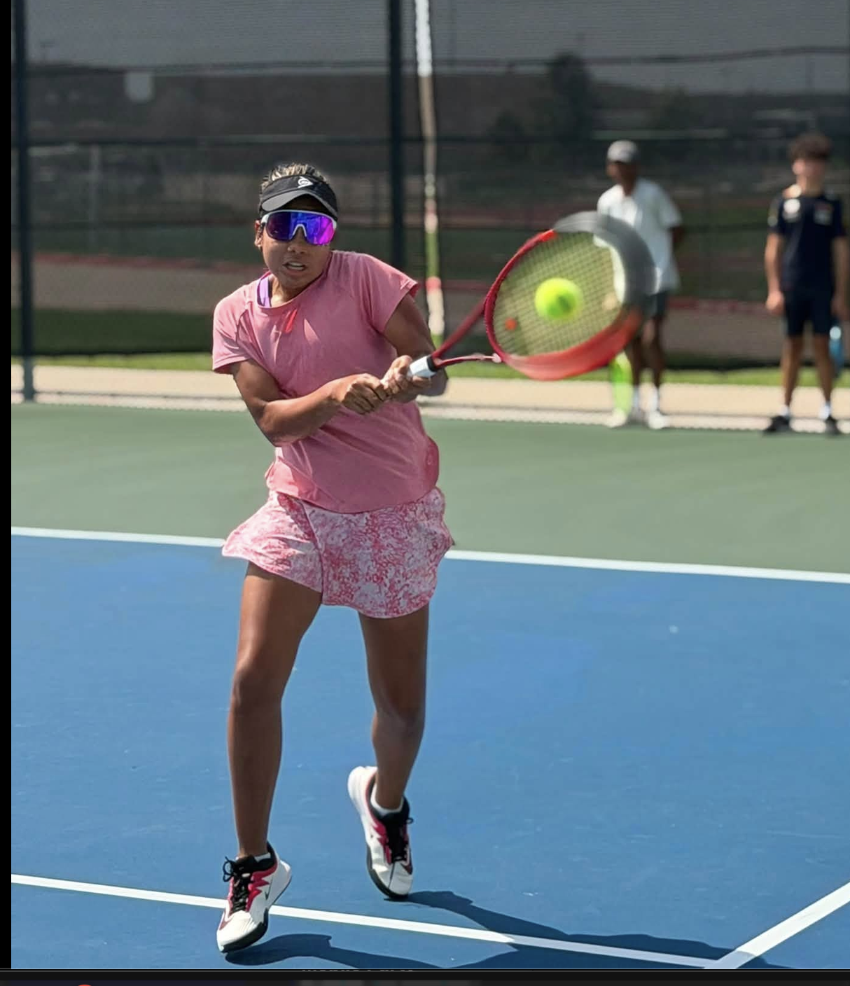 A woman playing tennis on a green court, wearing a pink outfit, black cap, and sunglasses, with the sun casting shadows.