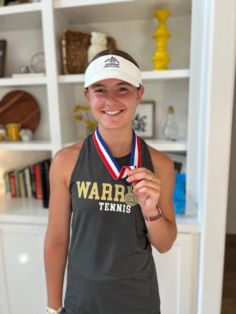 A young woman smiling, wearing a gray tennis tank top with 'WARRIORS TENNIS' printed on it, a white visor, and holding a silver medal on a red, white, and blue ribbon.