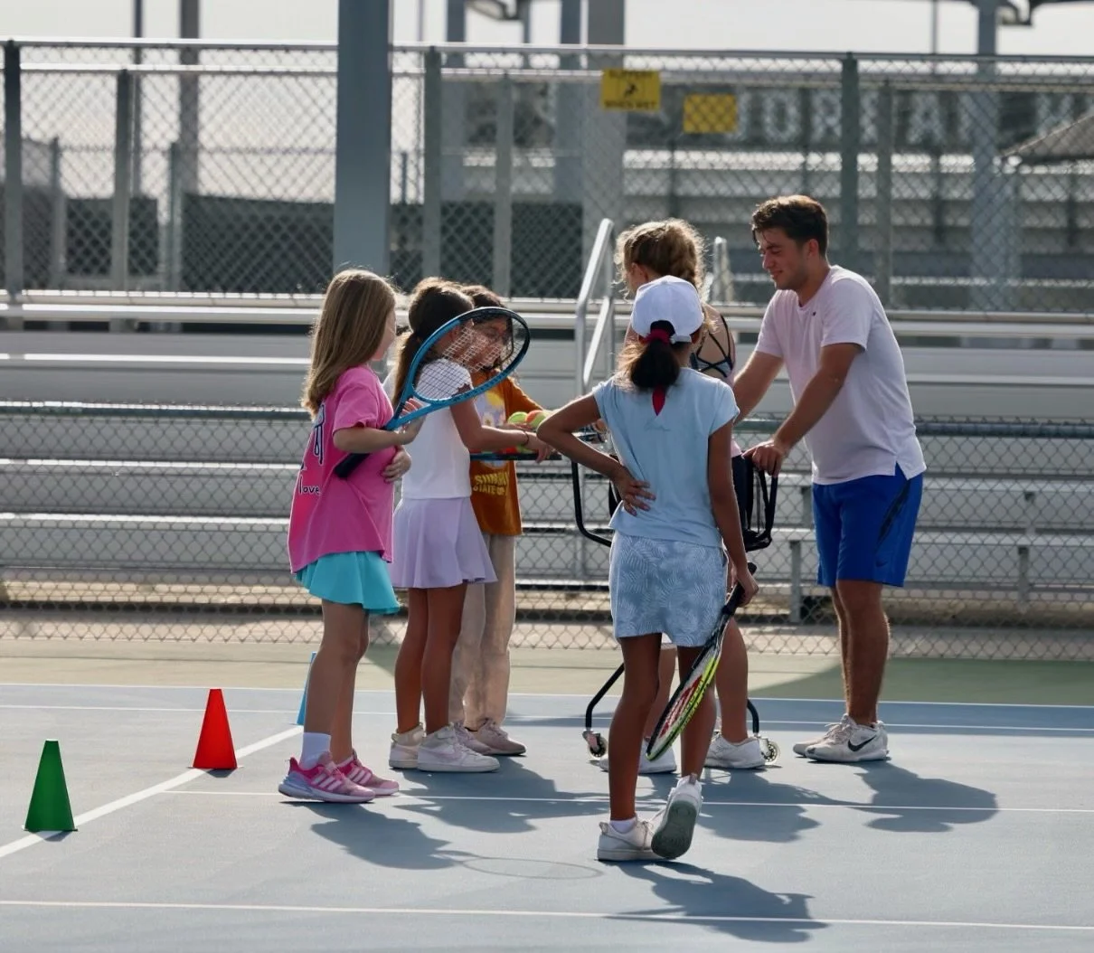 A young tennis coach giving instructions to a group of children holding tennis rackets on an outdoor tennis court, with a chain-link fence and bleachers in the background.