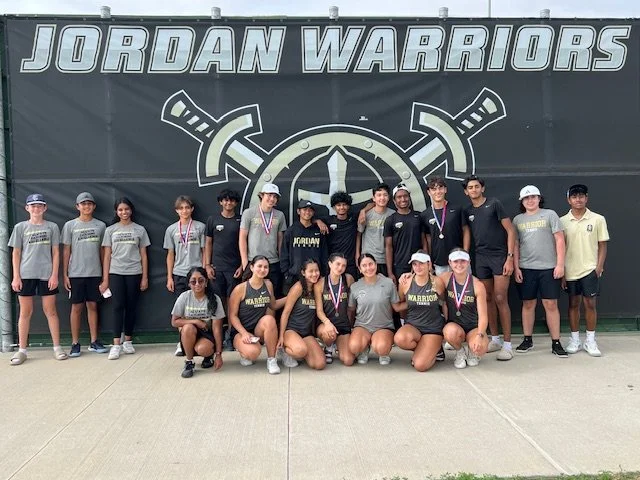 Group of young athletes and coaches in front of a large wall with 'Jordan Warriors' and a sword logo, posing for a team photo after a competition, some wearing medals.