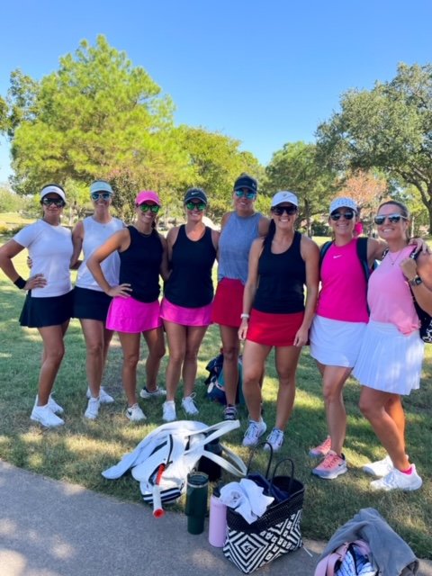 A group of eight women dressed in athletic clothing standing outdoors on a sunny day, with trees and grass in the background.