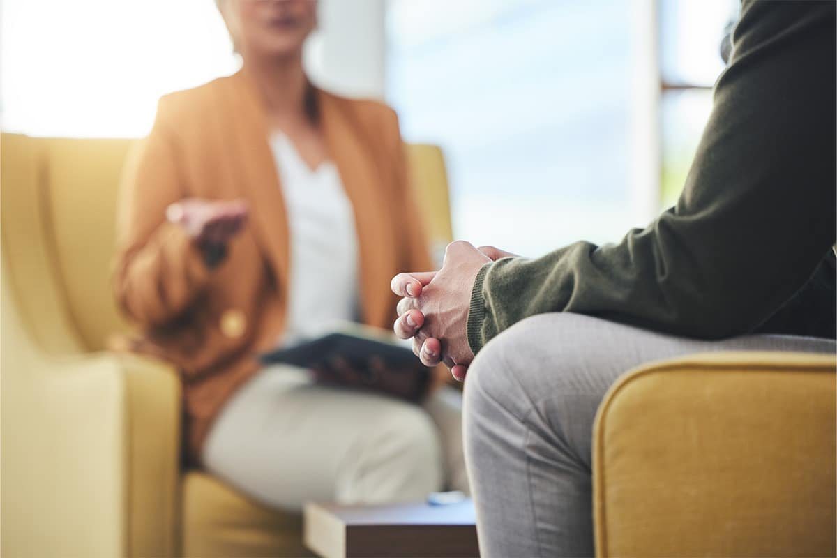 A person in a brown blazer sitting on a yellow couch during a therapy session, with hands clasped, with a therapist holding a mobile phone in the background.