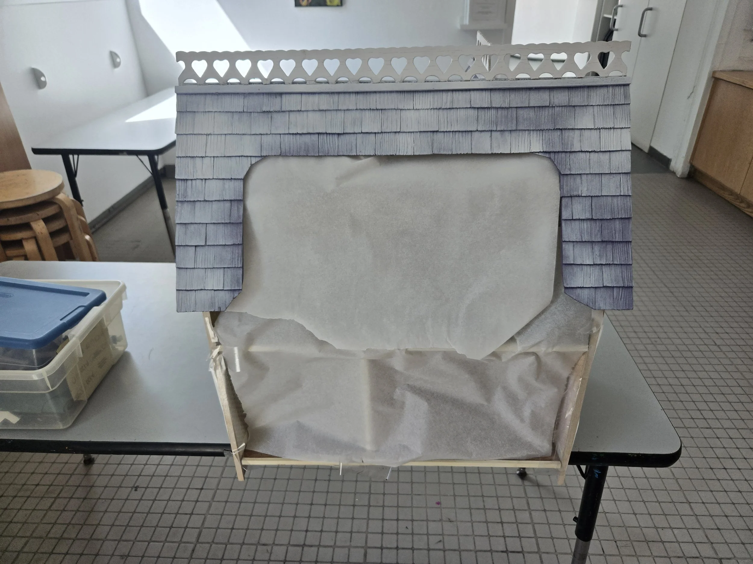 A miniature house model with gray shingles and a white decorative fence on top, placed on a table in a room with various furniture and storage containers in the background.