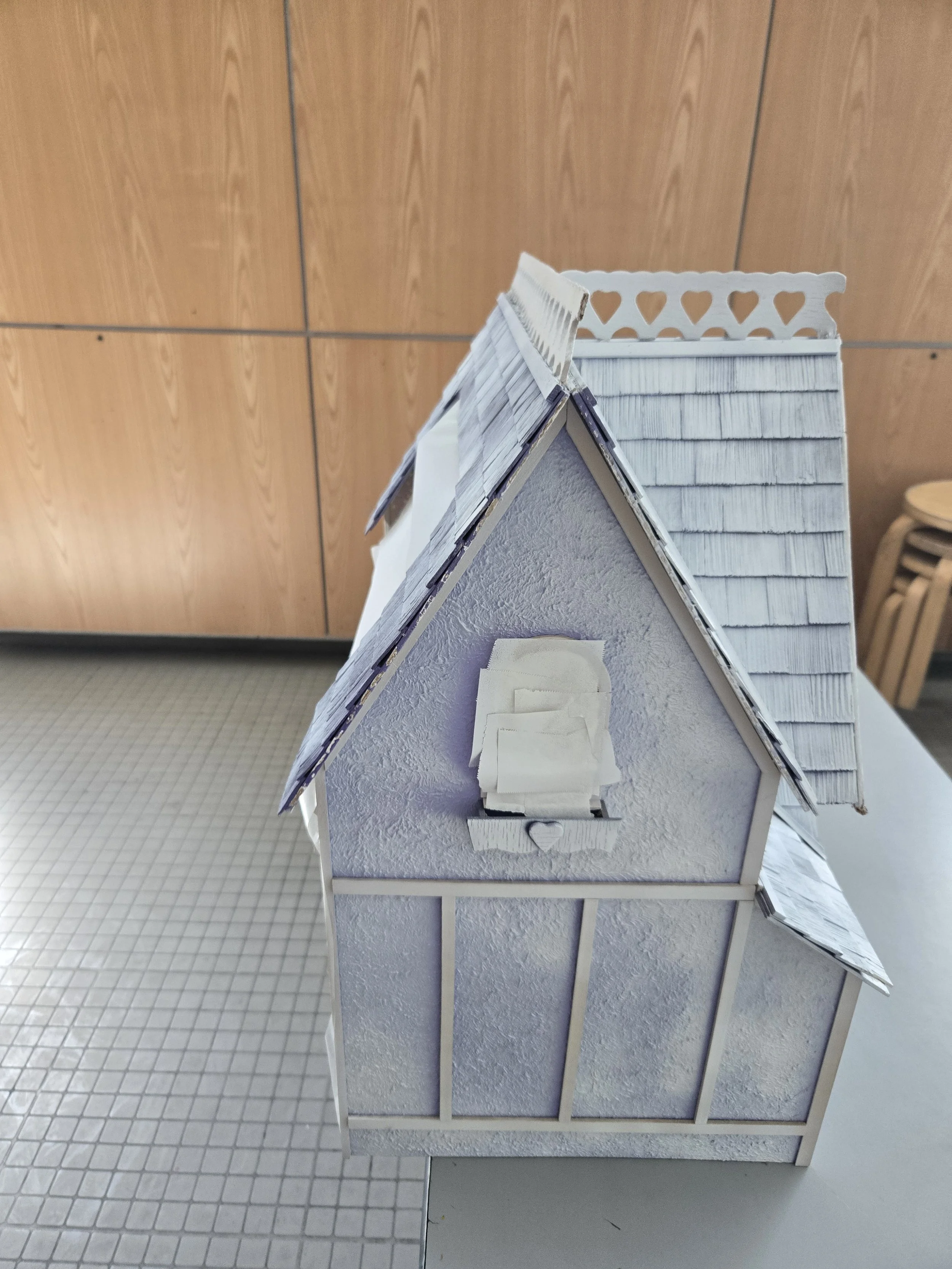 A small model house with a shingled roof, white textured walls, and a tiny front porch with a heart-shaped decoration, set against a wooden wall background.