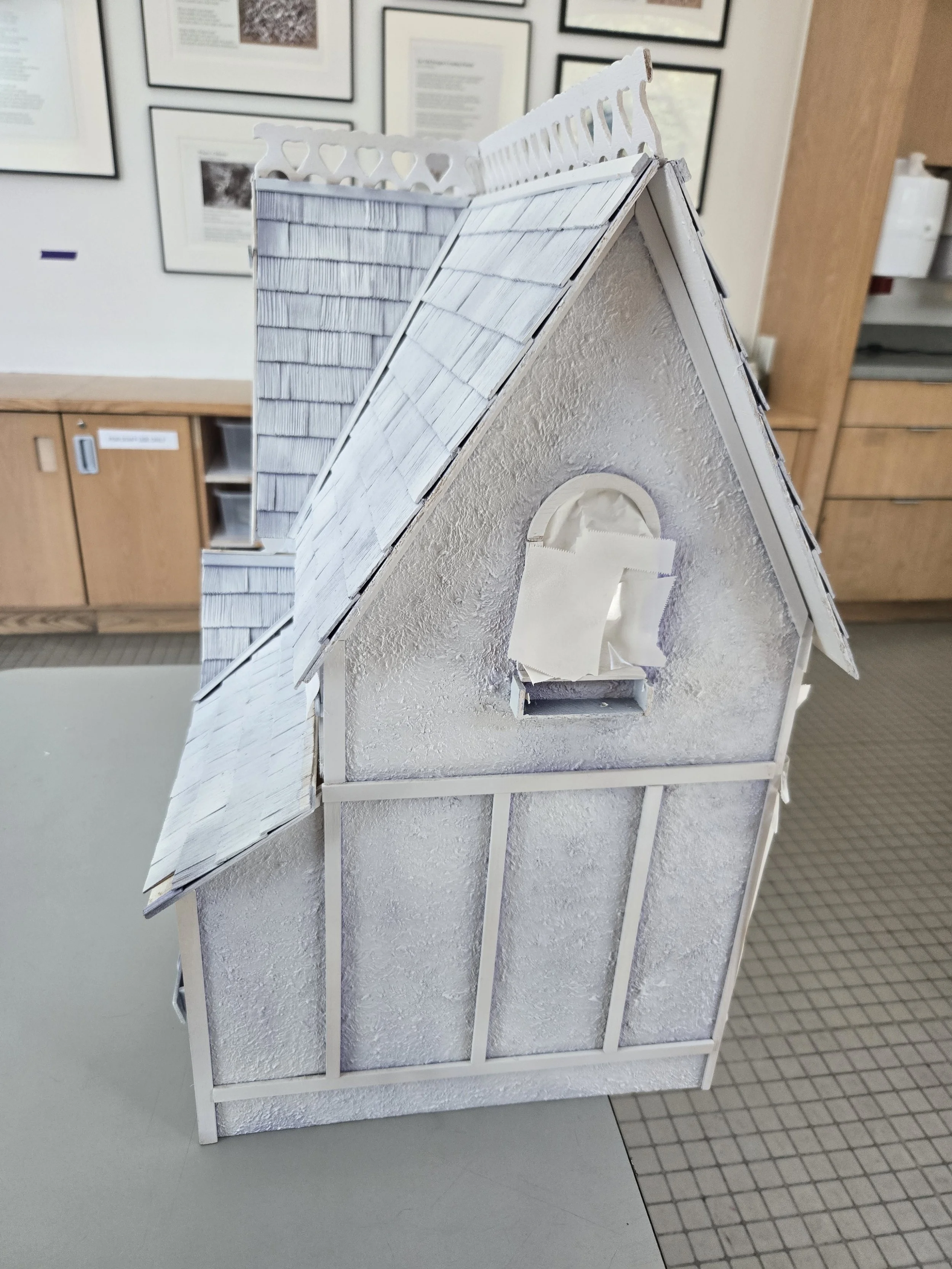 A miniature model of a house with a textured white wall, small shingle roof, and a tiny window with tissue paper curtains, displayed on a stand in an indoor setting with framed pictures on the wall.