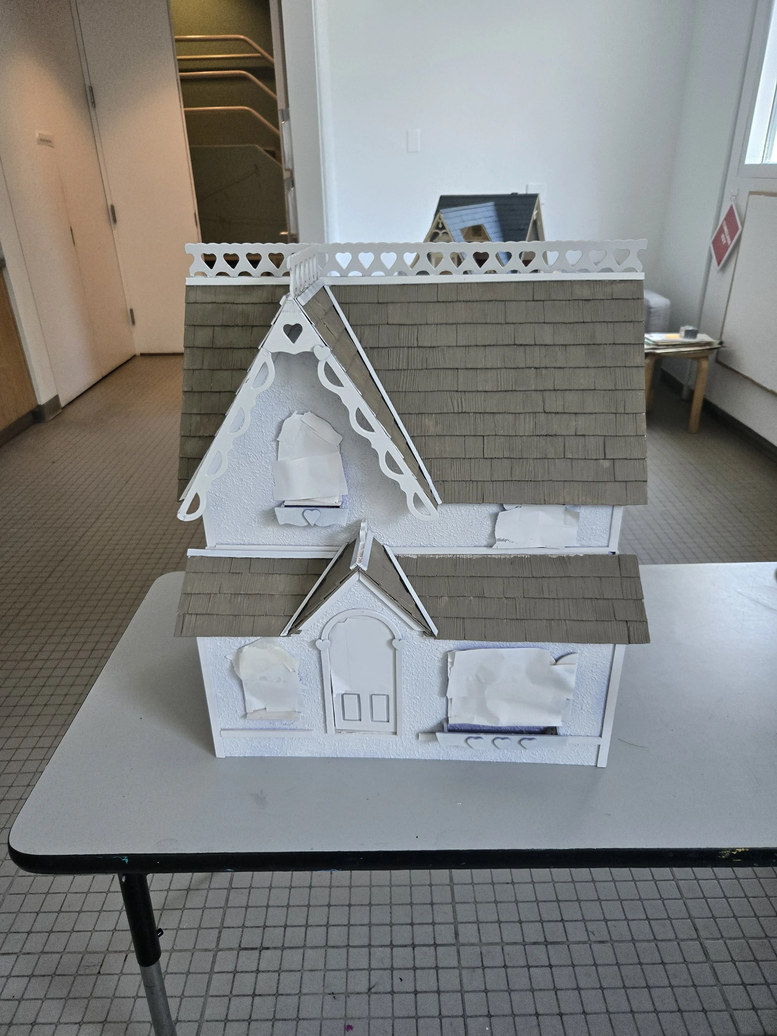This image shows a detailed model of a house placed on a table in a room. The model includes a white facade with windows and a door, and a roof made of brown shingles. Behind the model, there is a real house roof and a small house model with a dark b